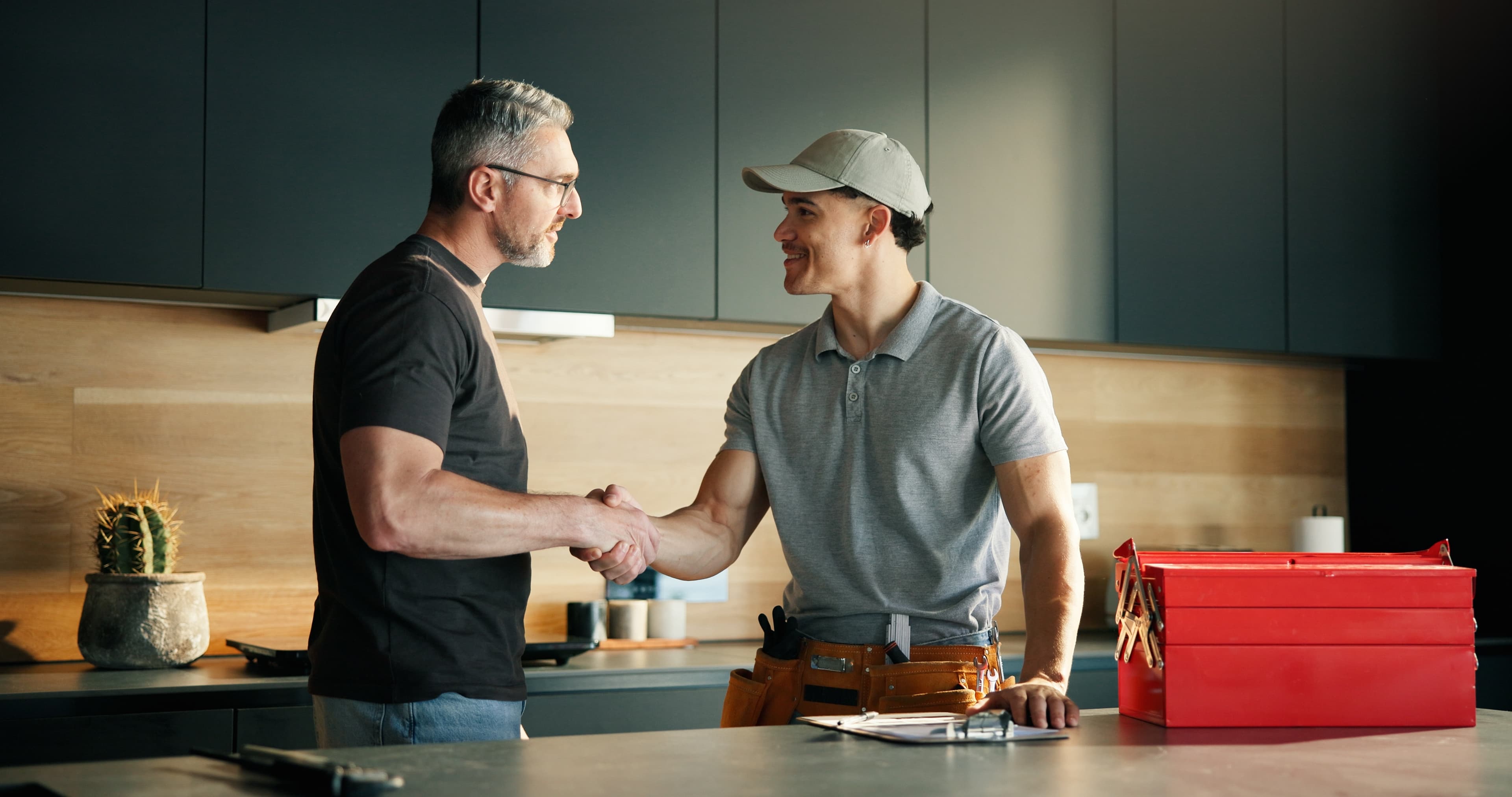 Two men shaking hands in a kitchen beside a clipboard and a red toolbox on the counter