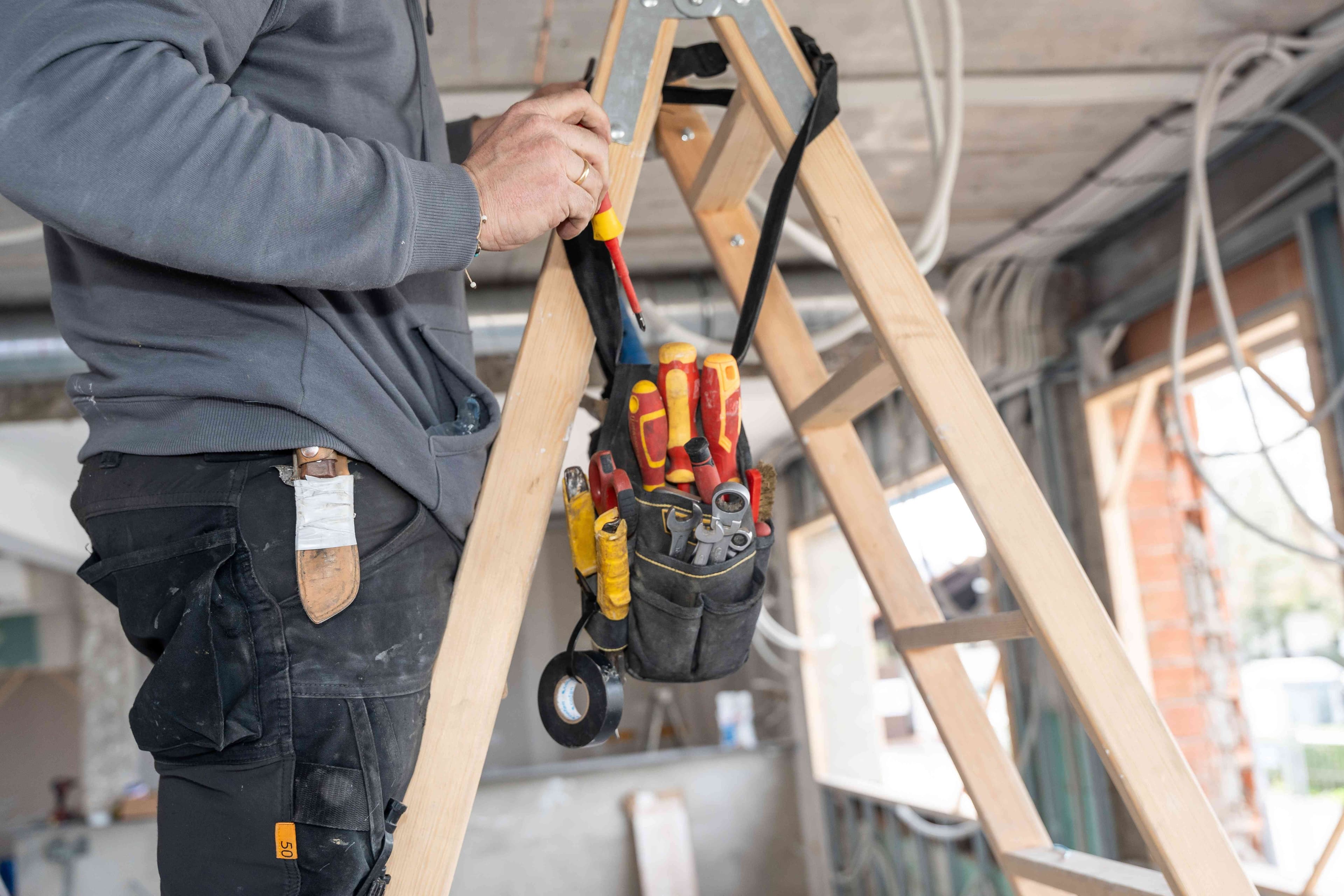 Close-up of a hand holding a screwdriver near a wooden ladder with a tool pouch holding screwdrivers, wrenches, and tape