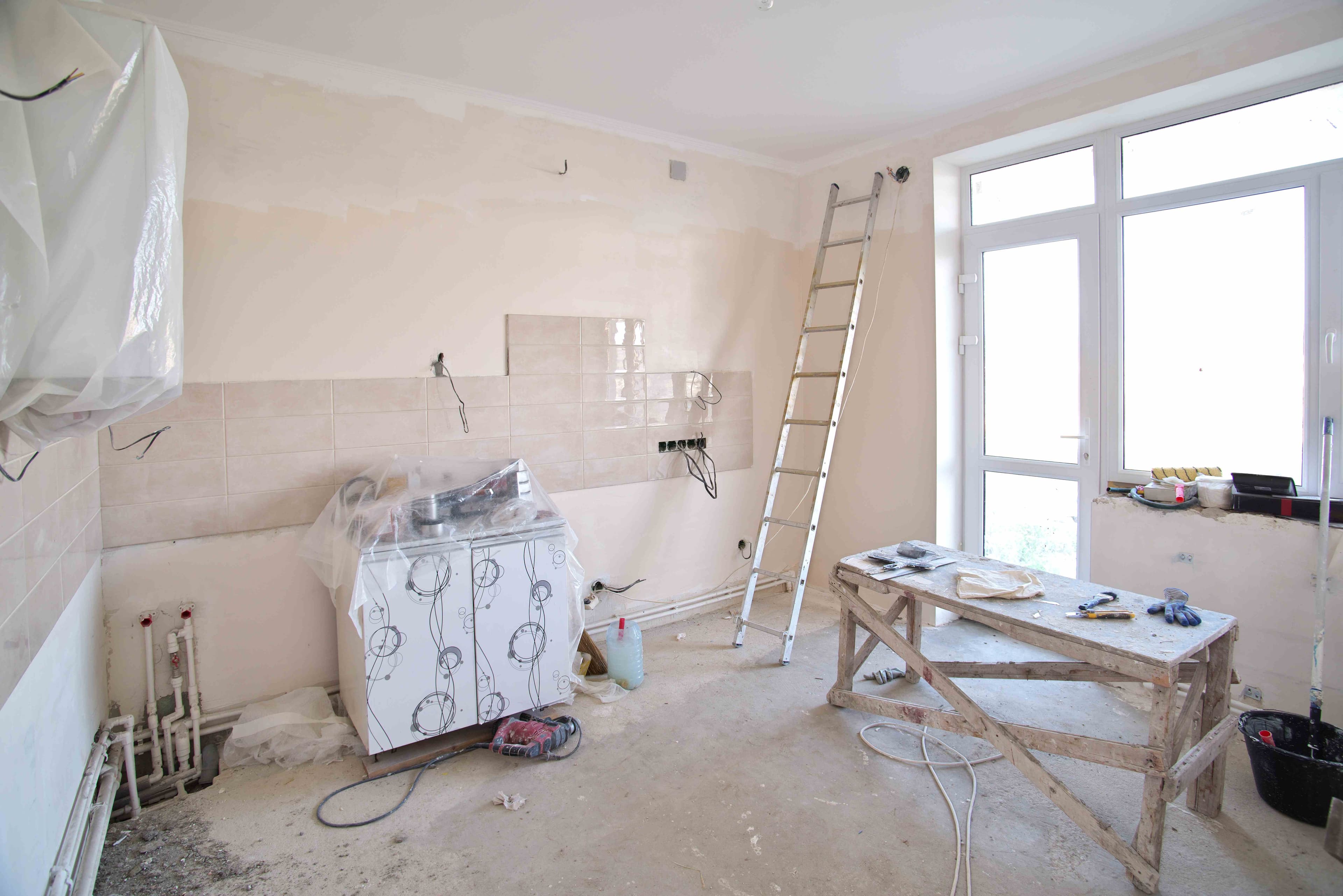 Kitchen under renovation with a ladder, workbench, tools, and exposed plumbing and wiring