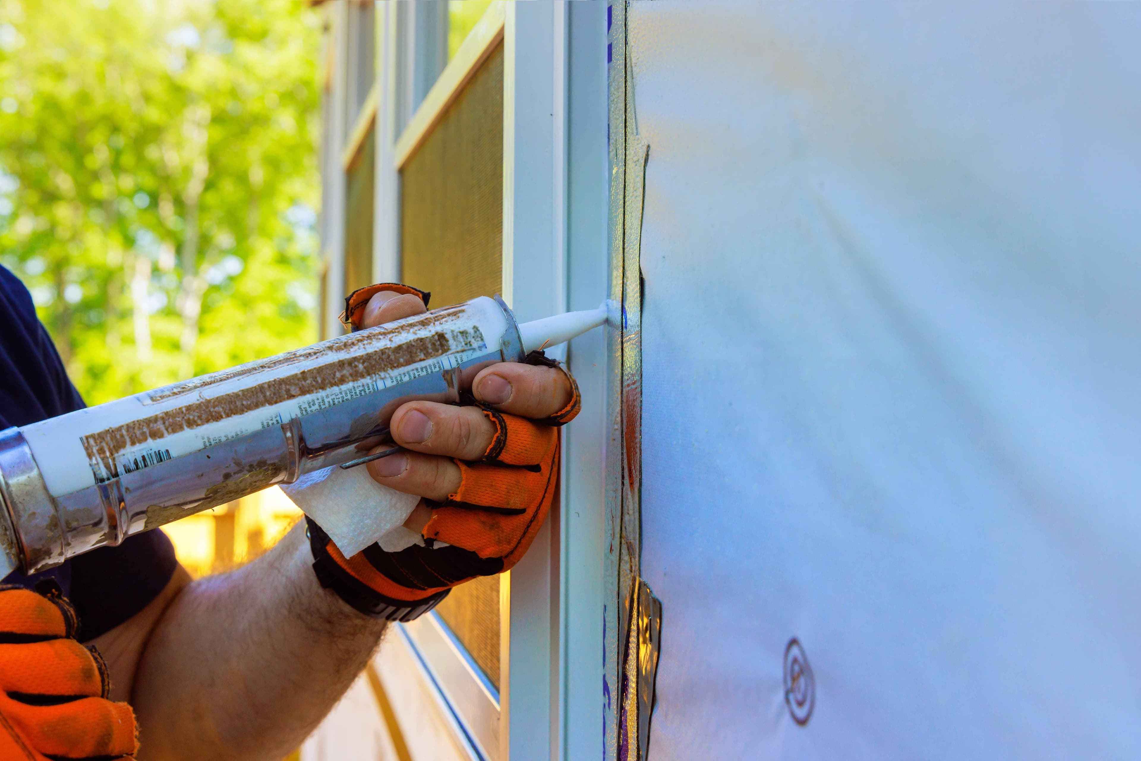 A gloved hand applying caulk along the joint between a window frame and exterior siding