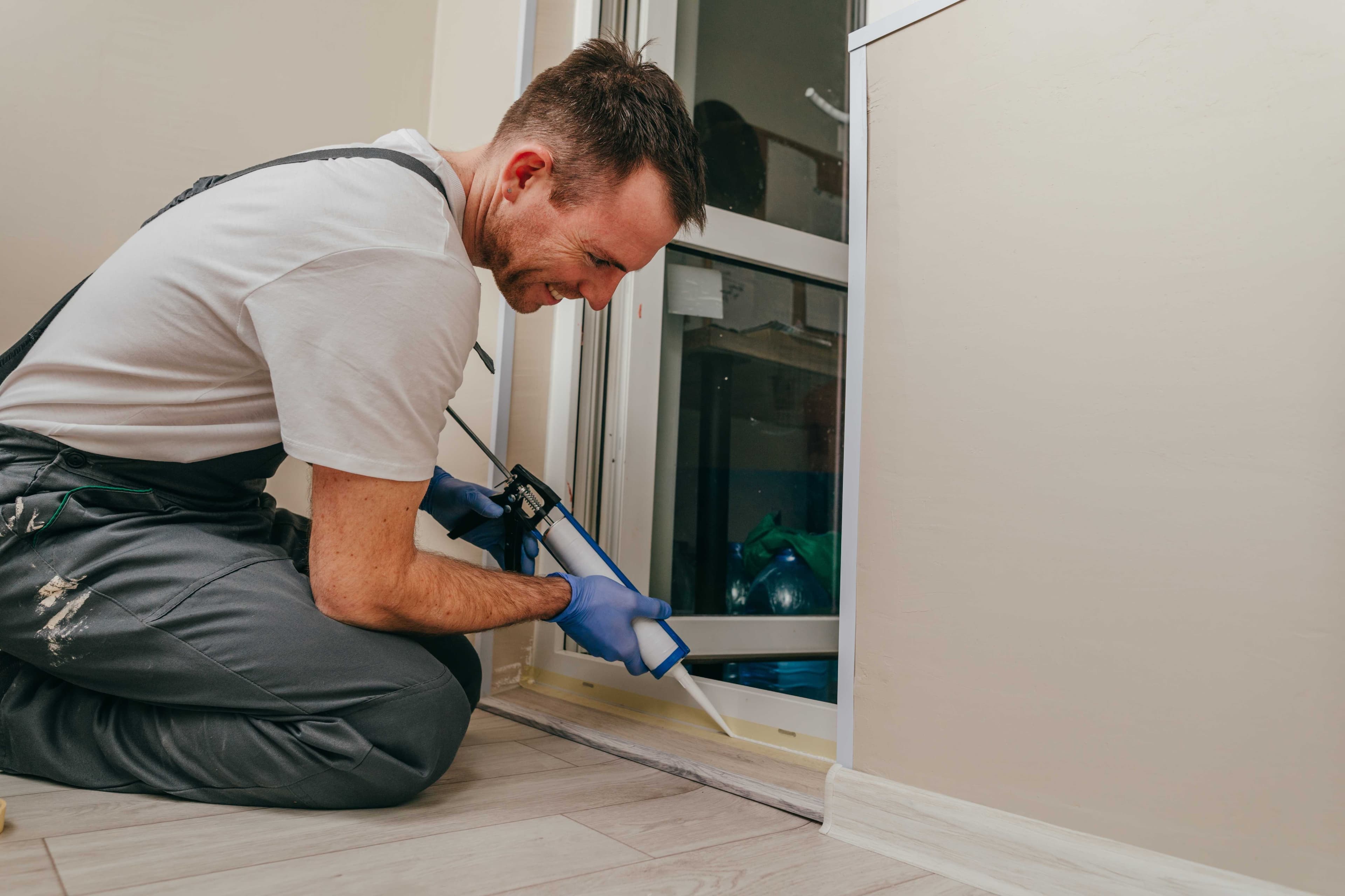 A worker kneeling to apply caulk along a sliding door threshold during a home repair