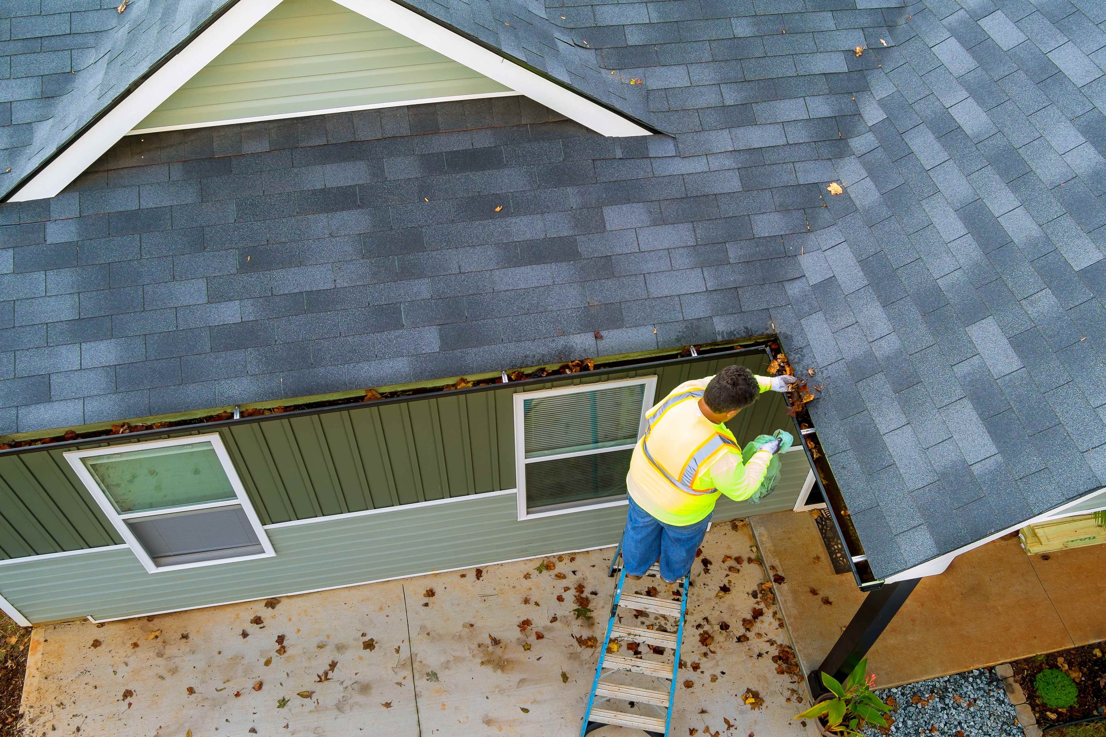 Aerial view of a worker in a high-visibility vest standing on a ladder cleaning leaves from a gutter along a two-story home's roofline