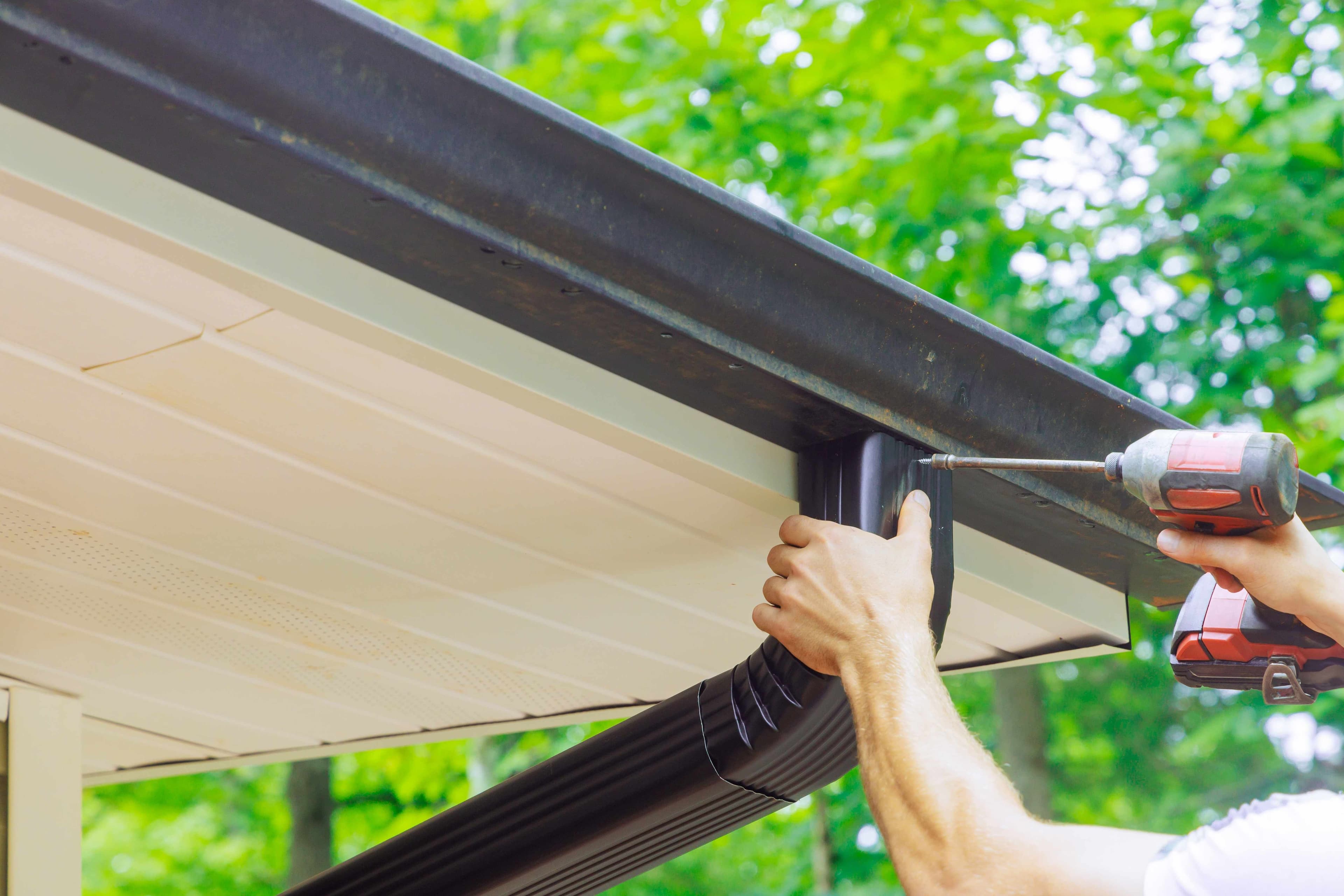 Hands using a power drill to attach a black downspout bracket beneath a metal roof eave surrounded by green trees