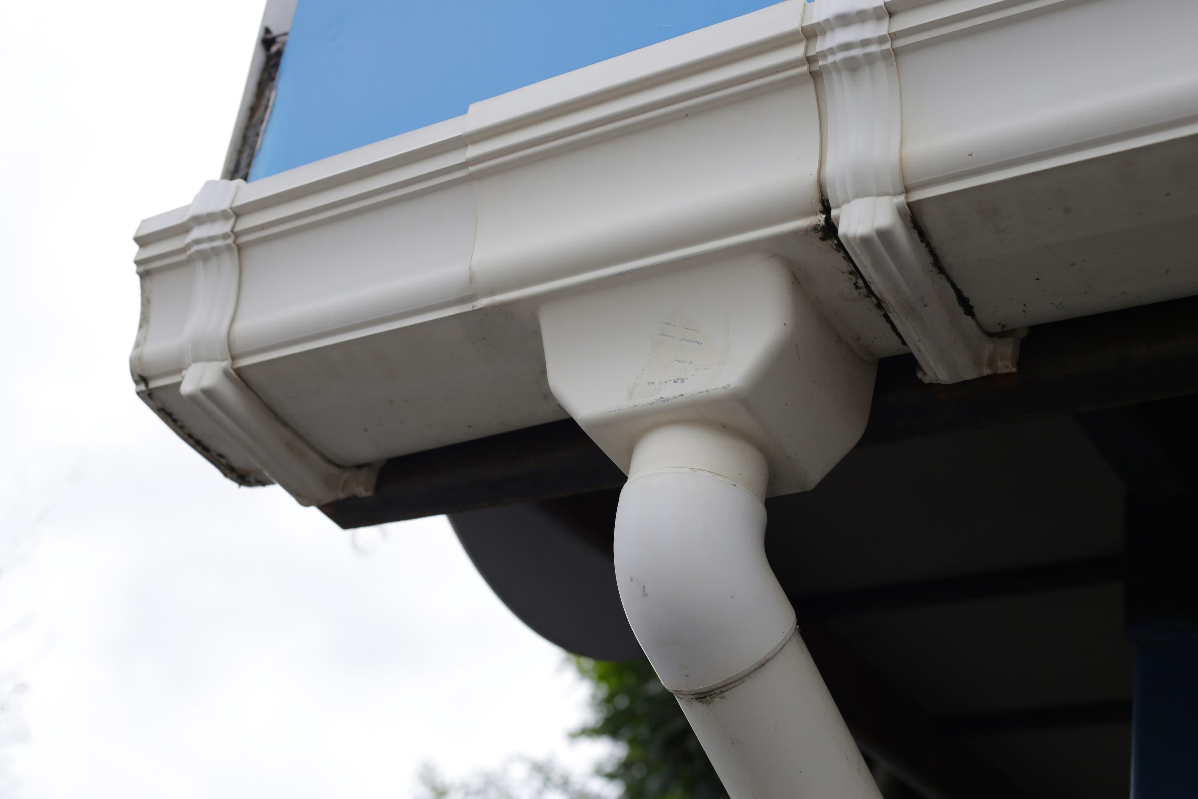 Close-up view of a white gutter corner piece and downspout outlet attached beneath a home's roofline against a blue sky