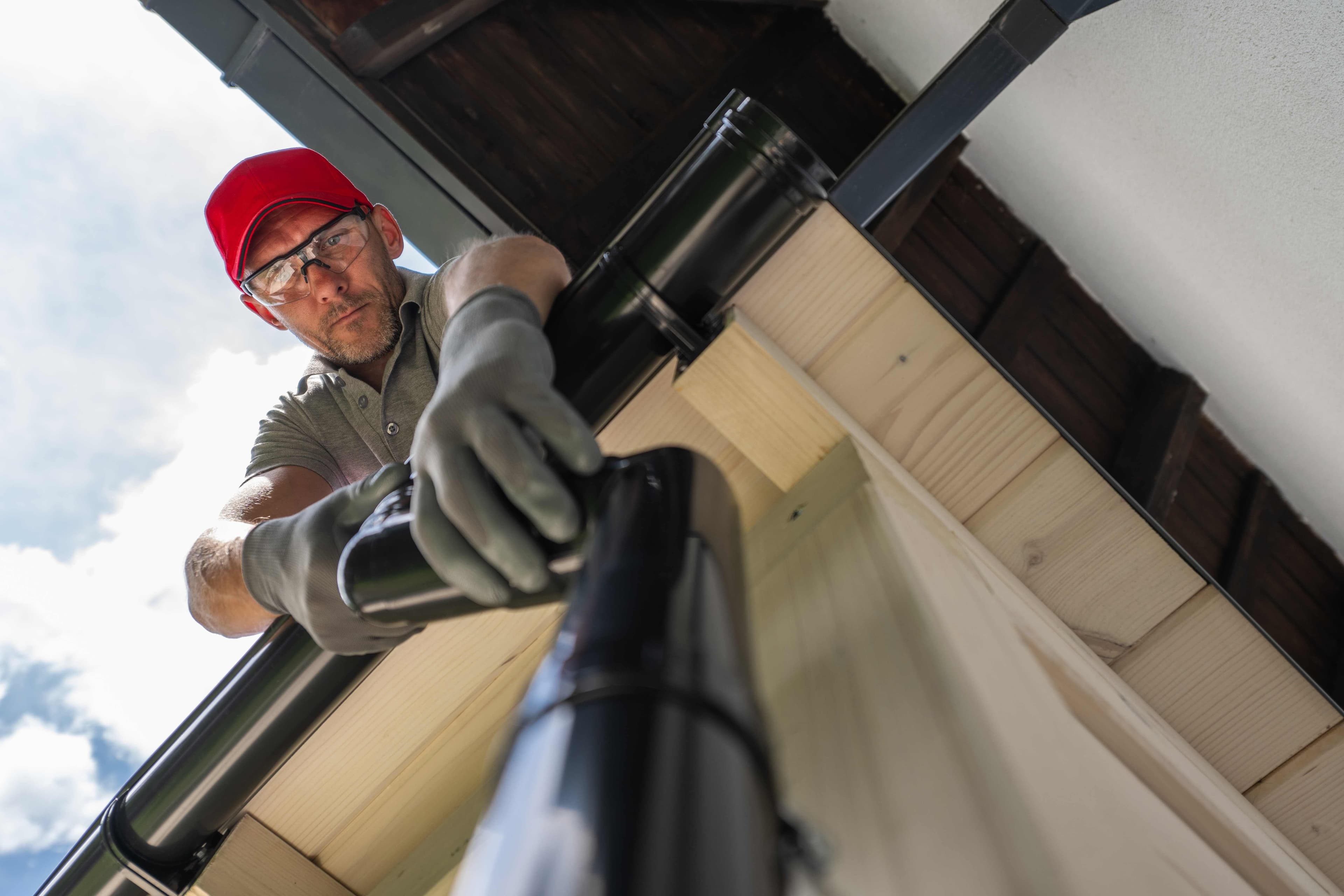 A man wearing a red cap and safety glasses closely inspecting a black downspout pipe joint beneath a wood roof eave