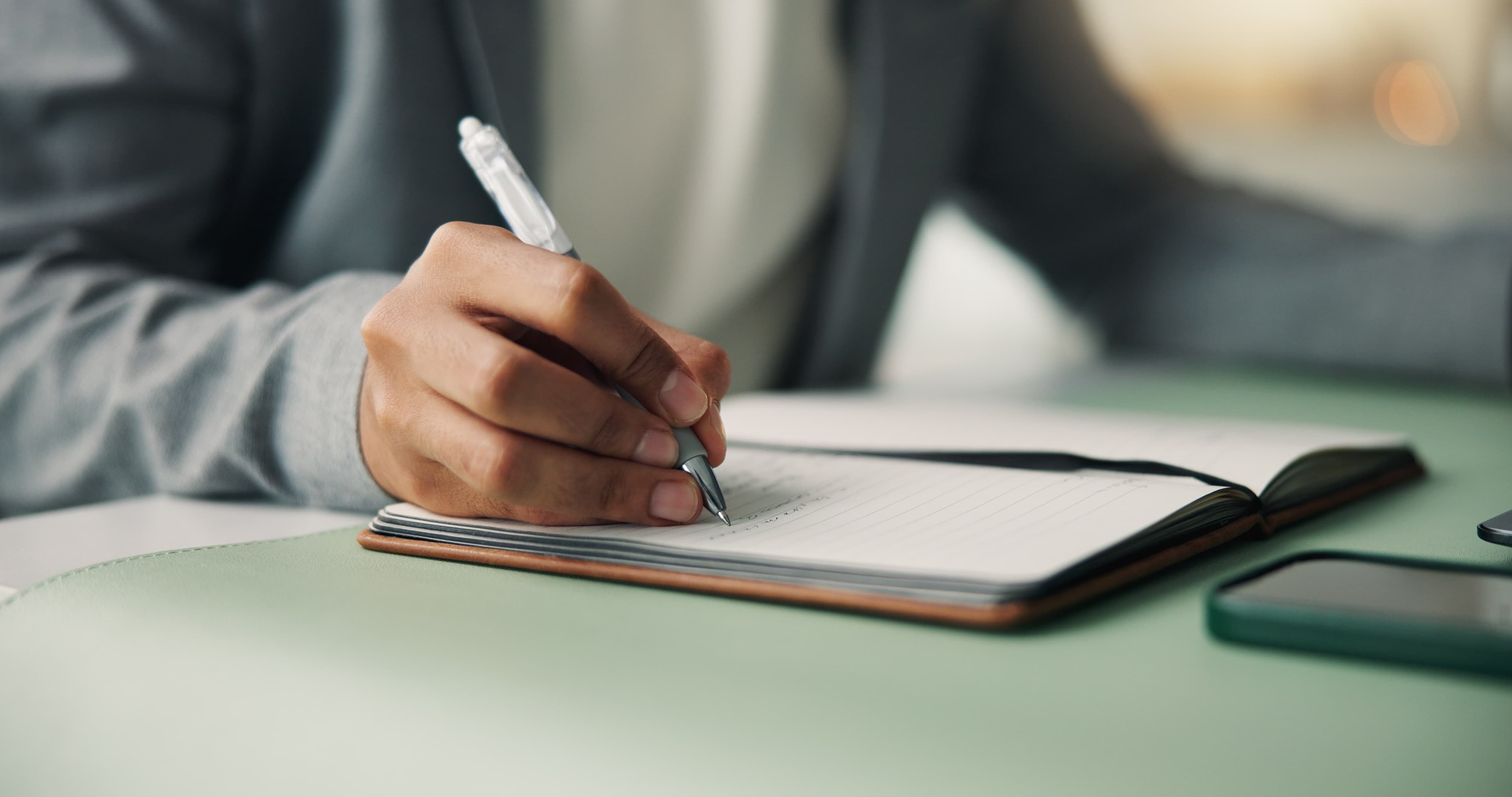 A close-up of a hand writing notes with a pen in a lined leather notebook on a desk, with a smartphone nearby.