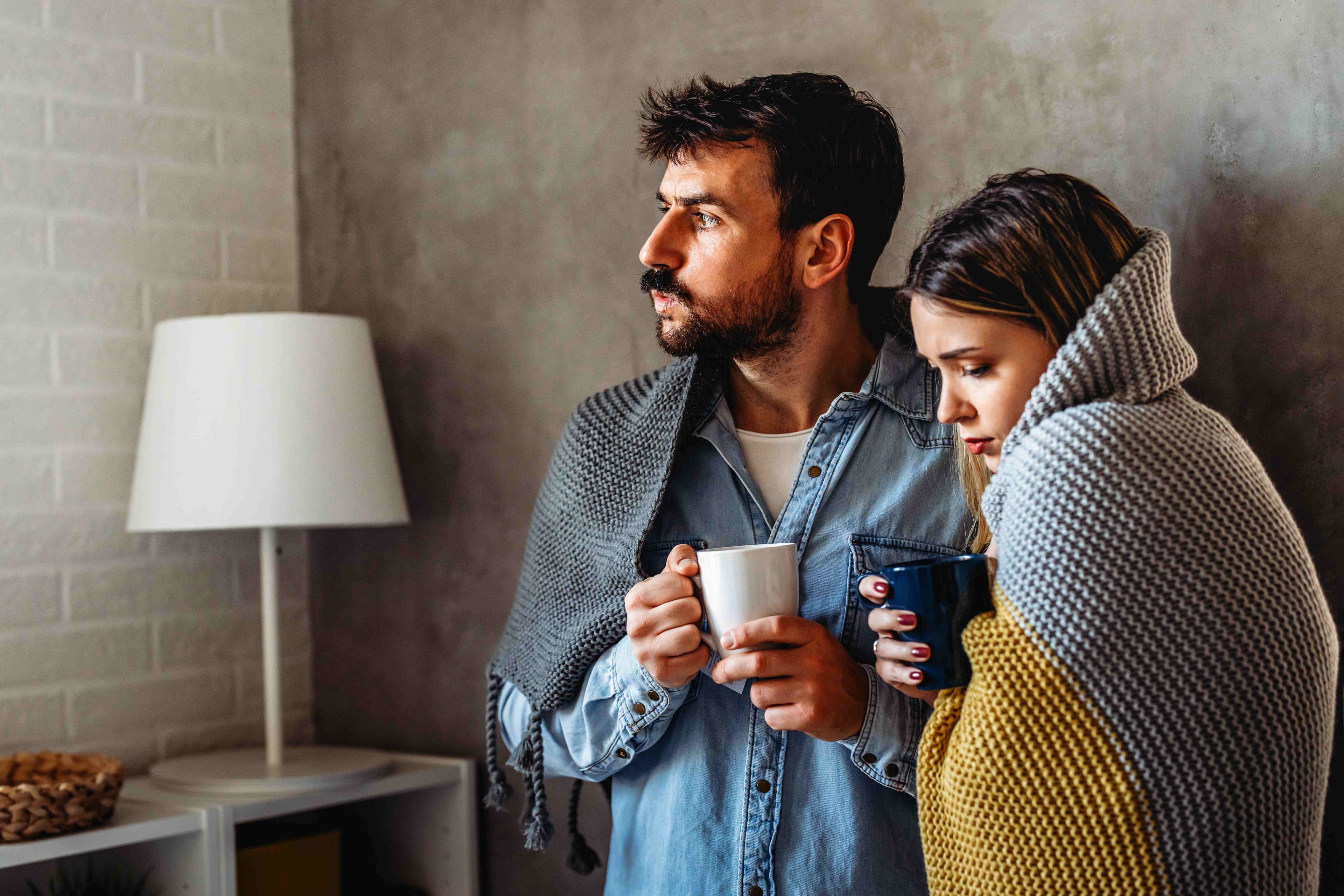 A couple wrapped in blankets holding warm mugs and looking out a window during cold weather