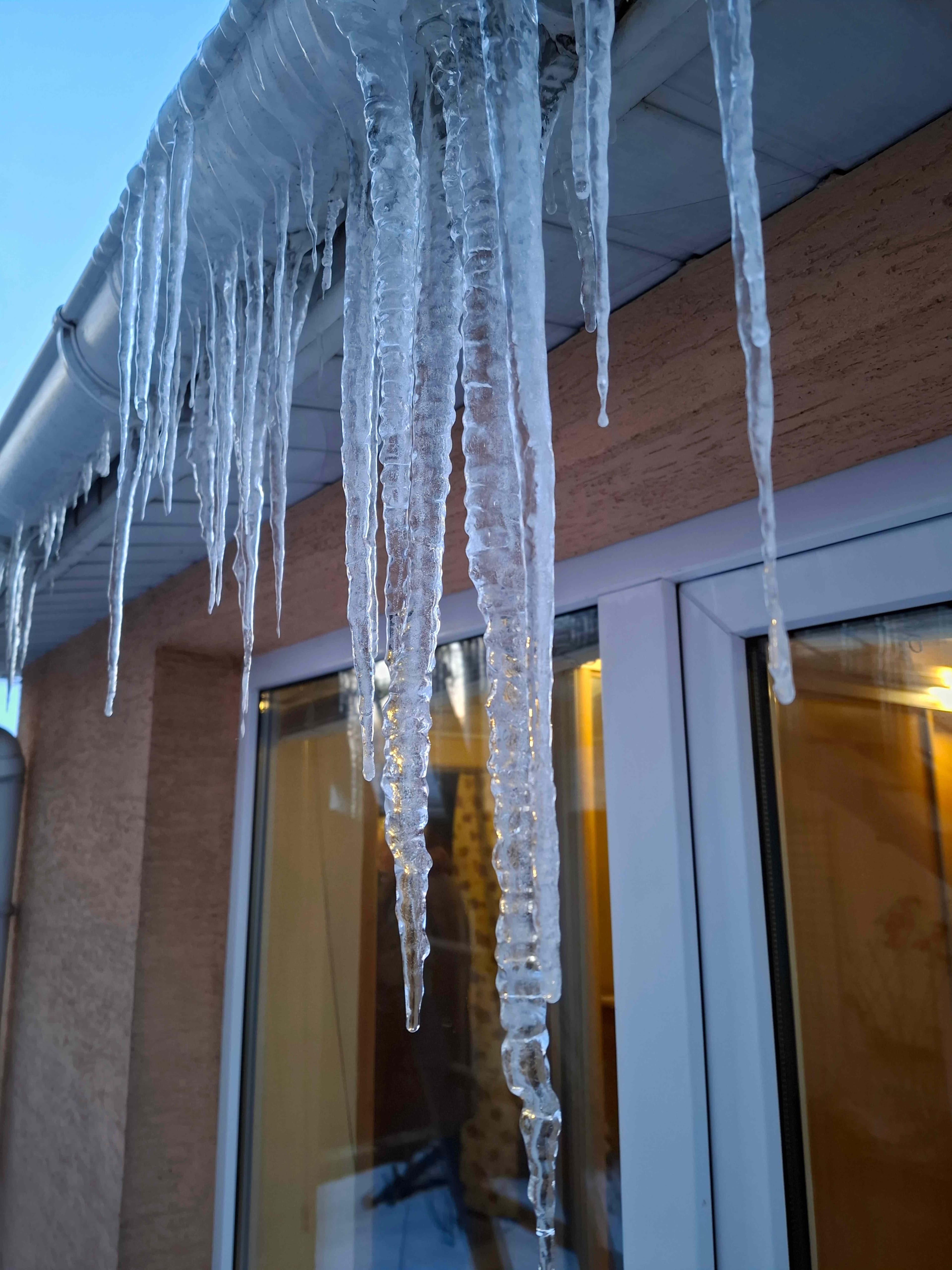 Icicles hanging from a roofline at dusk with warm light glowing from the windows behind them