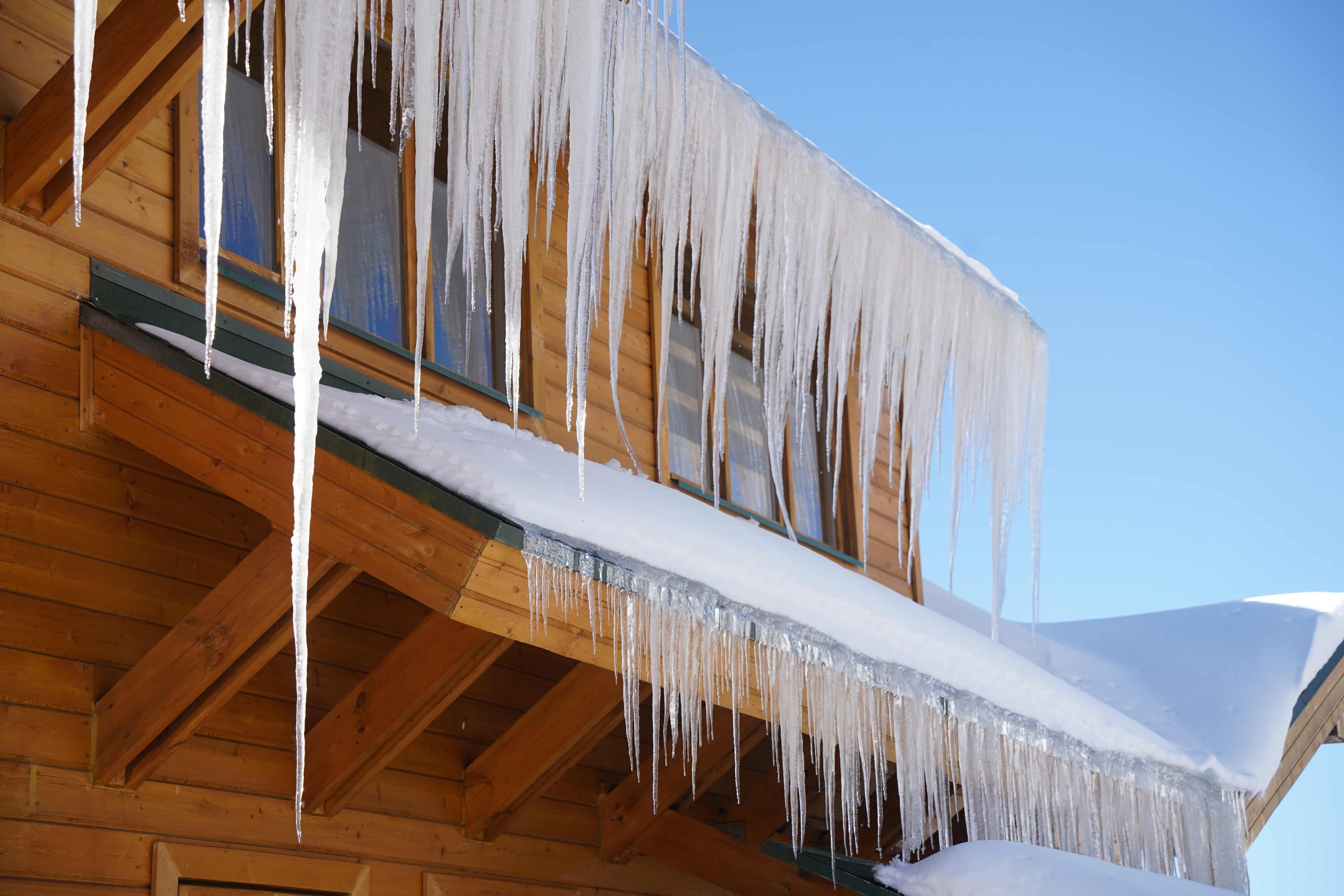 Icicles hanging from the snow-covered eaves of a wood-sided home under a clear blue sky