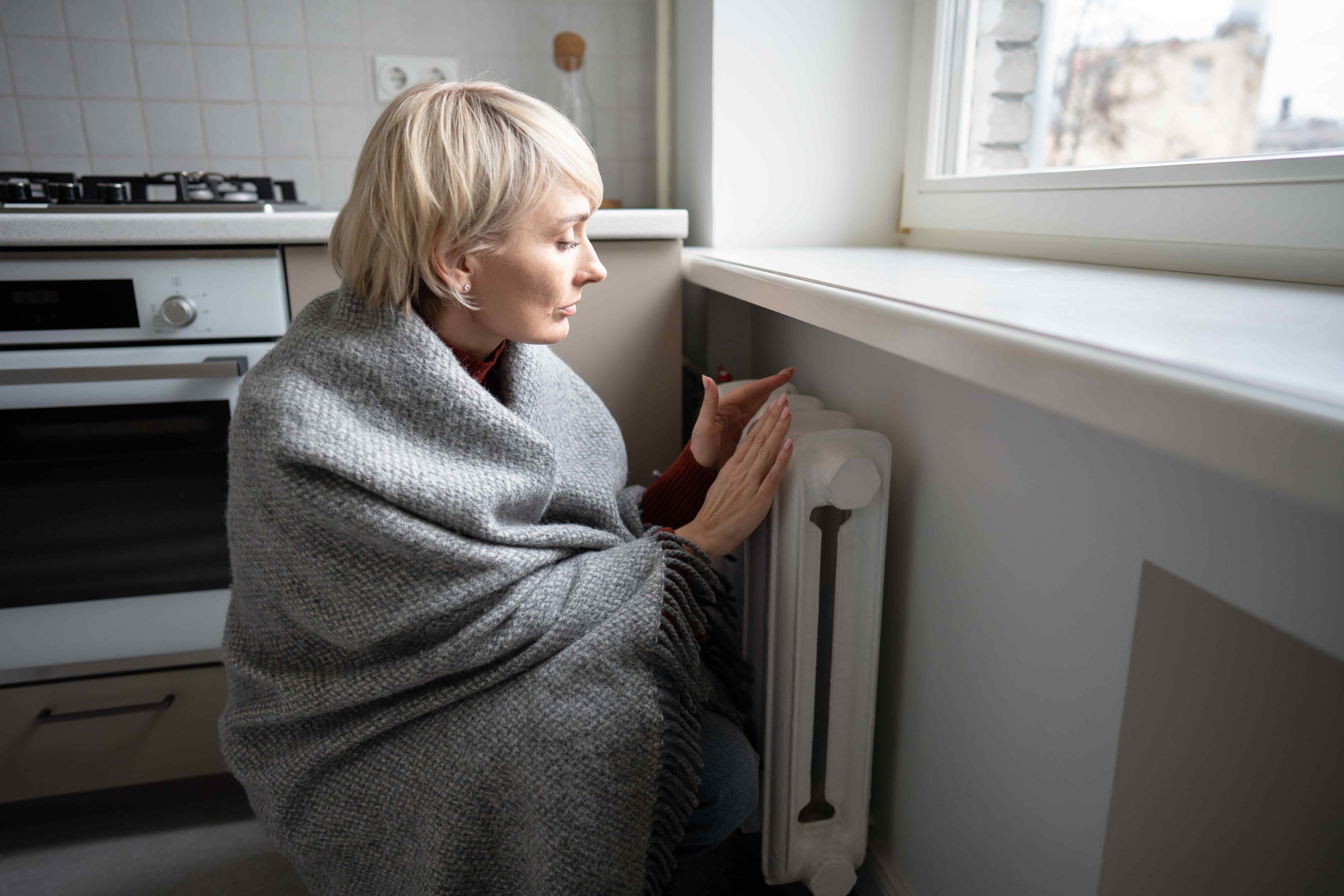 A woman wrapped in a blanket warming her hands near a radiator by a window