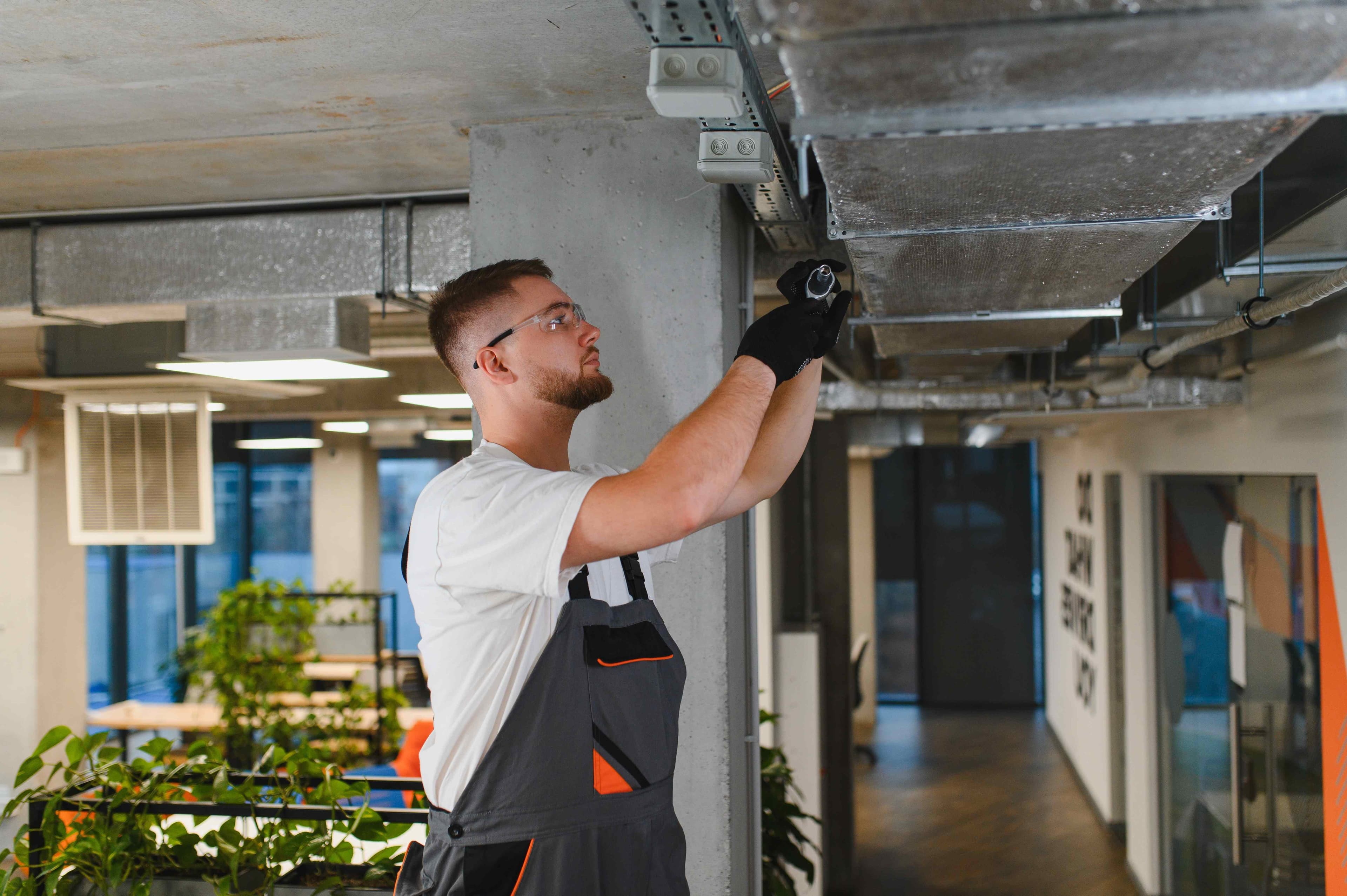 Worker in safety glasses and gloves adjusting a ceiling conduit box next to insulated ductwork in a modern office