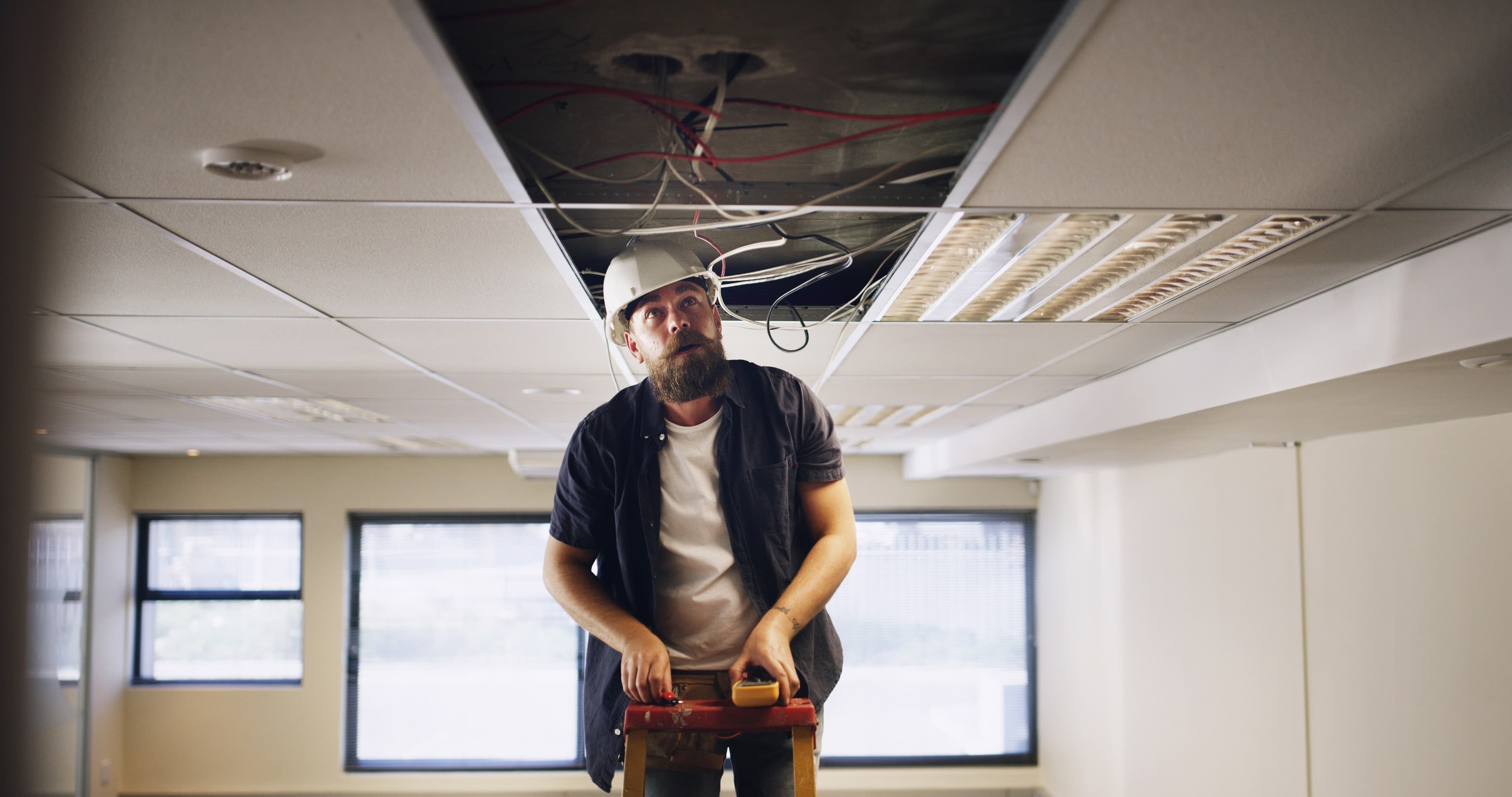 Worker wearing a hard hat and holding a multimeter while looking up at an open ceiling with exposed wiring