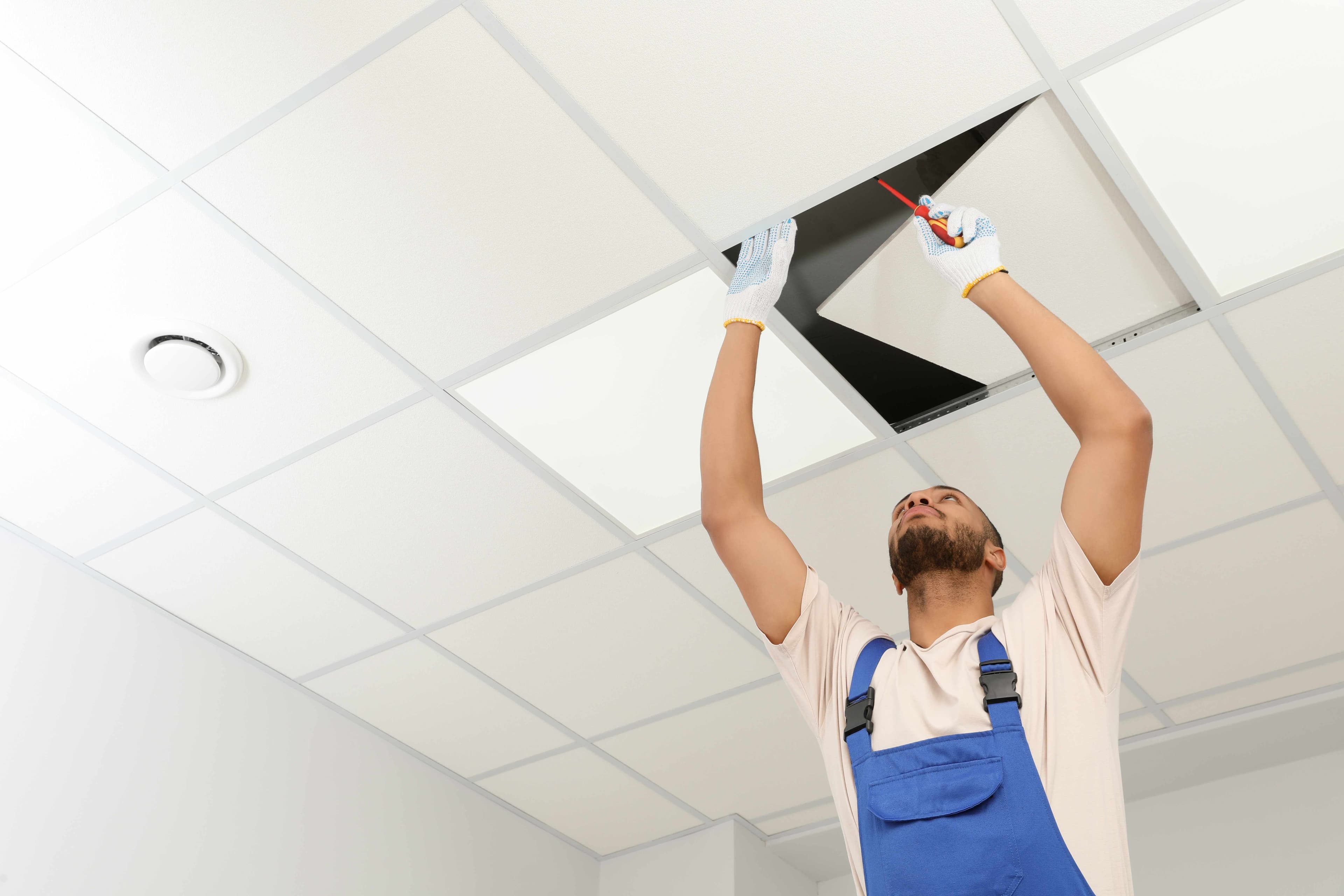 Worker in gloves lifting a suspended ceiling tile with a screwdriver in an office space