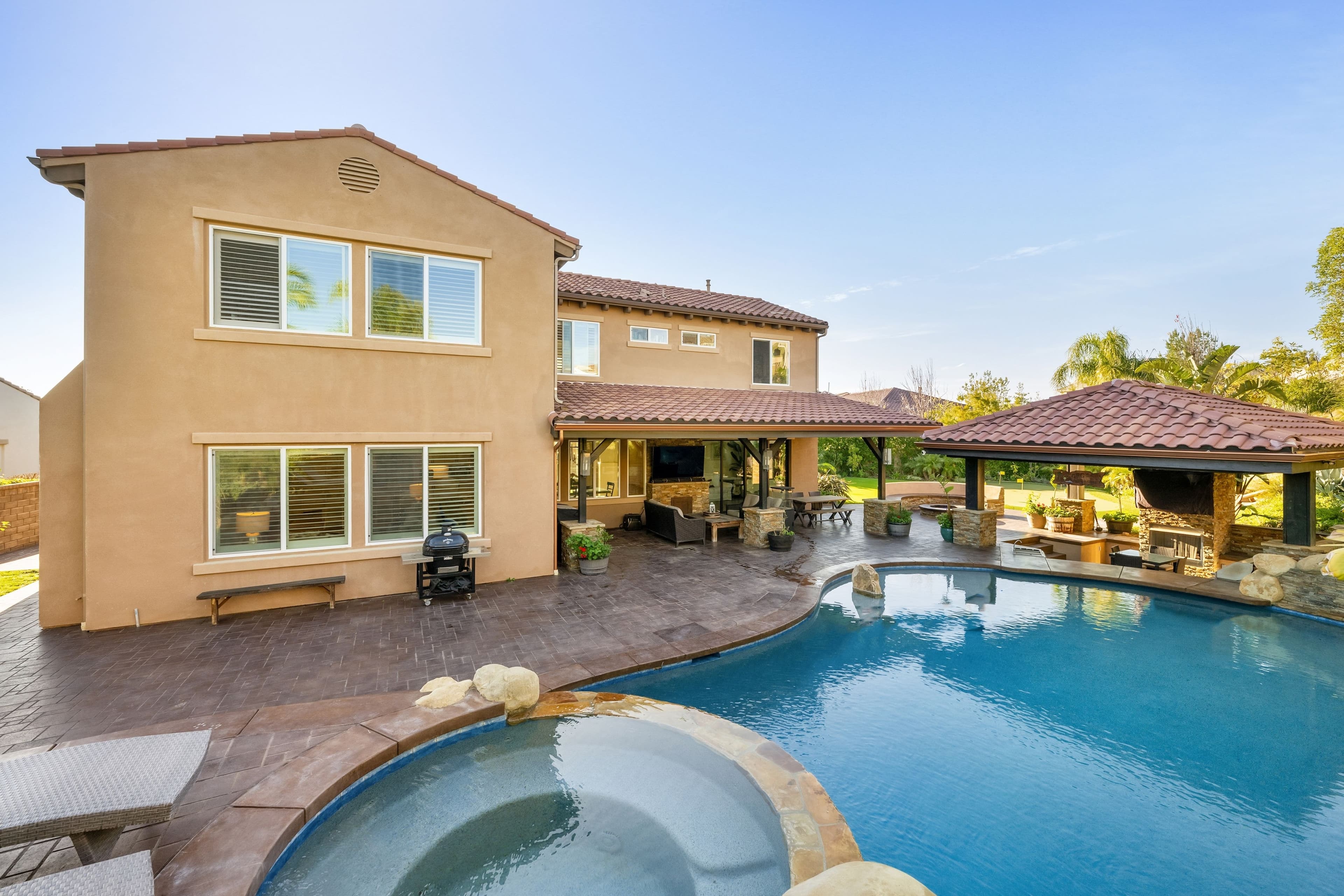 Two-story home with a covered patio, outdoor kitchen, and pergola overlooking a curved swimming pool and spa
