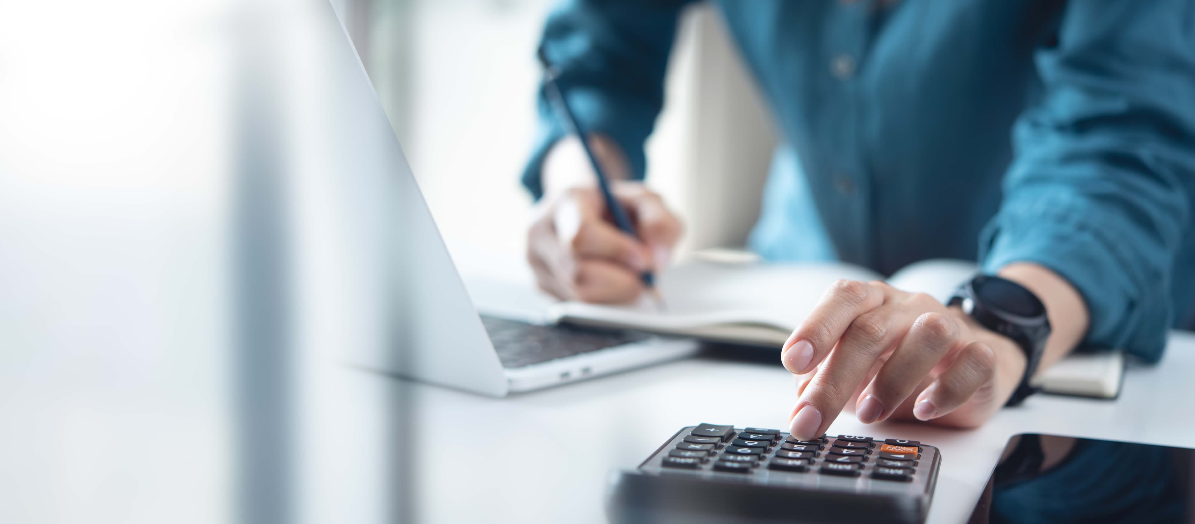 Close-up of a hand pressing keys on a calculator while the other hand writes notes beside an open laptop