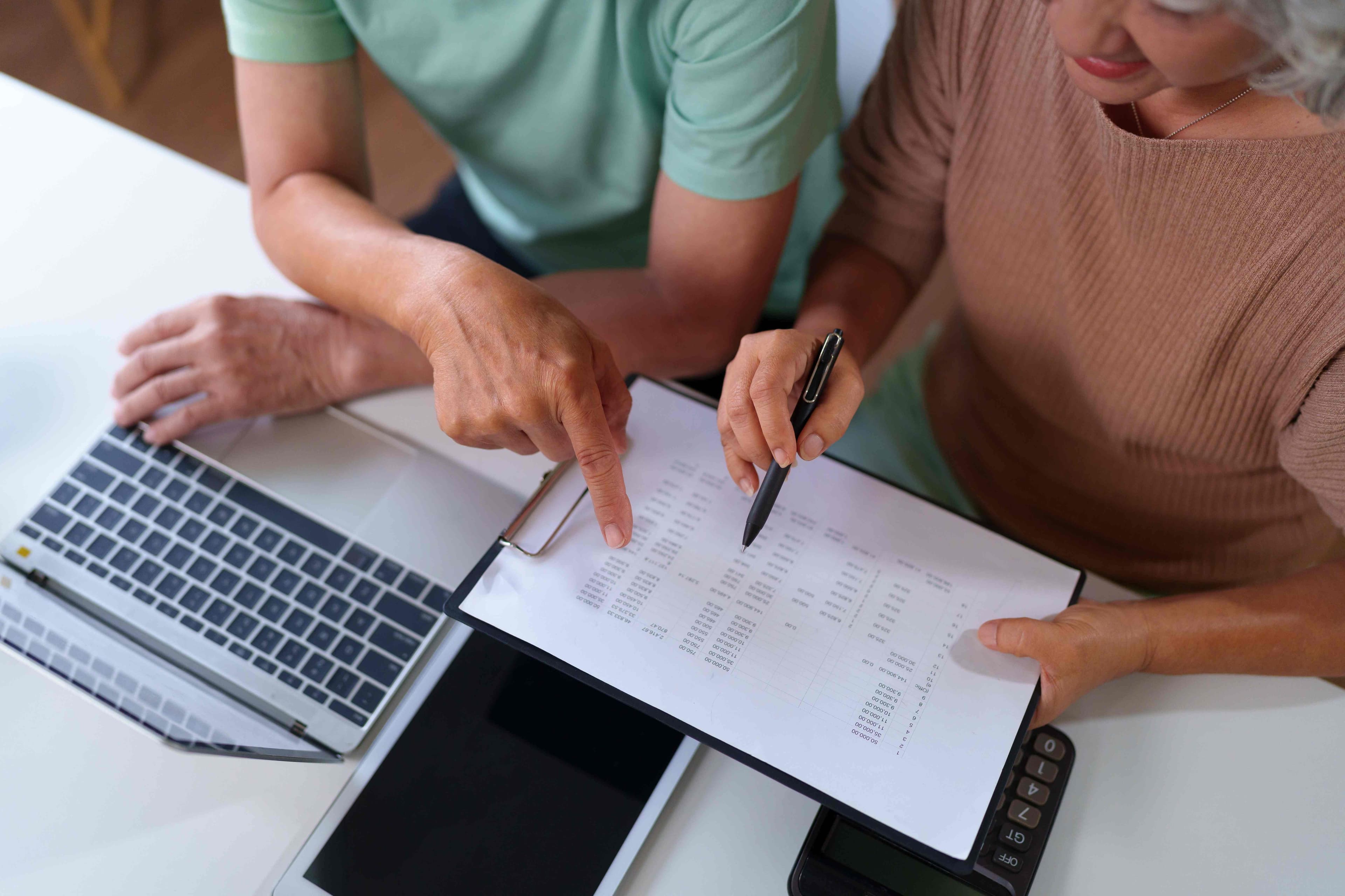 Two people reviewing a printed spreadsheet of figures on a clipboard next to an open laptop