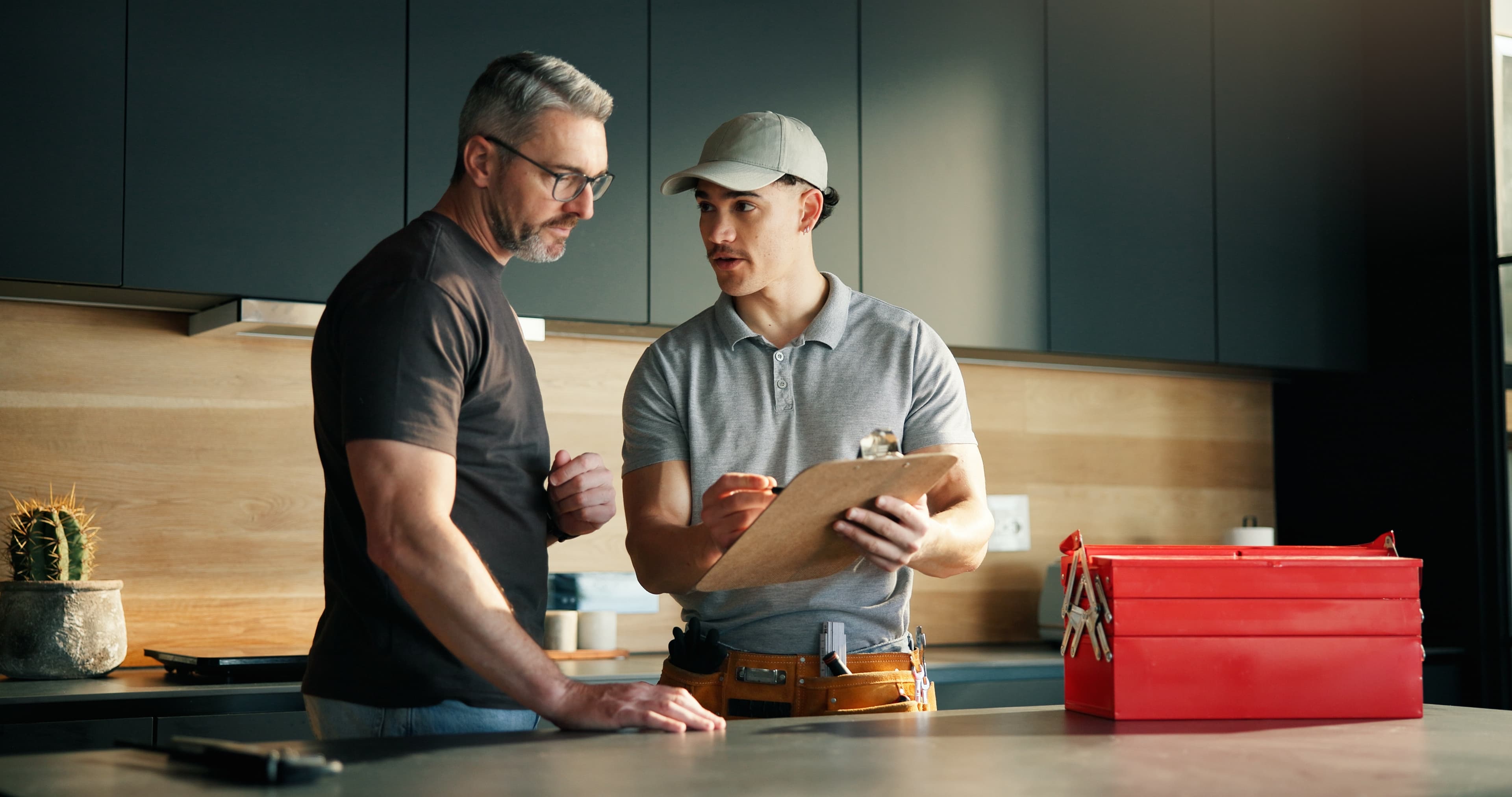 Two people talking in a kitchen while one holds a clipboard, with a red toolbox on the counter nearby
