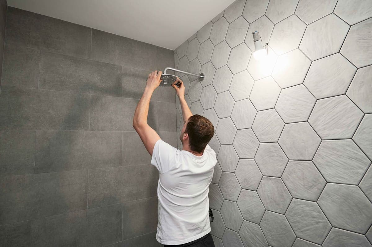 Person adjusting a rain-style shower head in a bathroom with gray hexagon tile