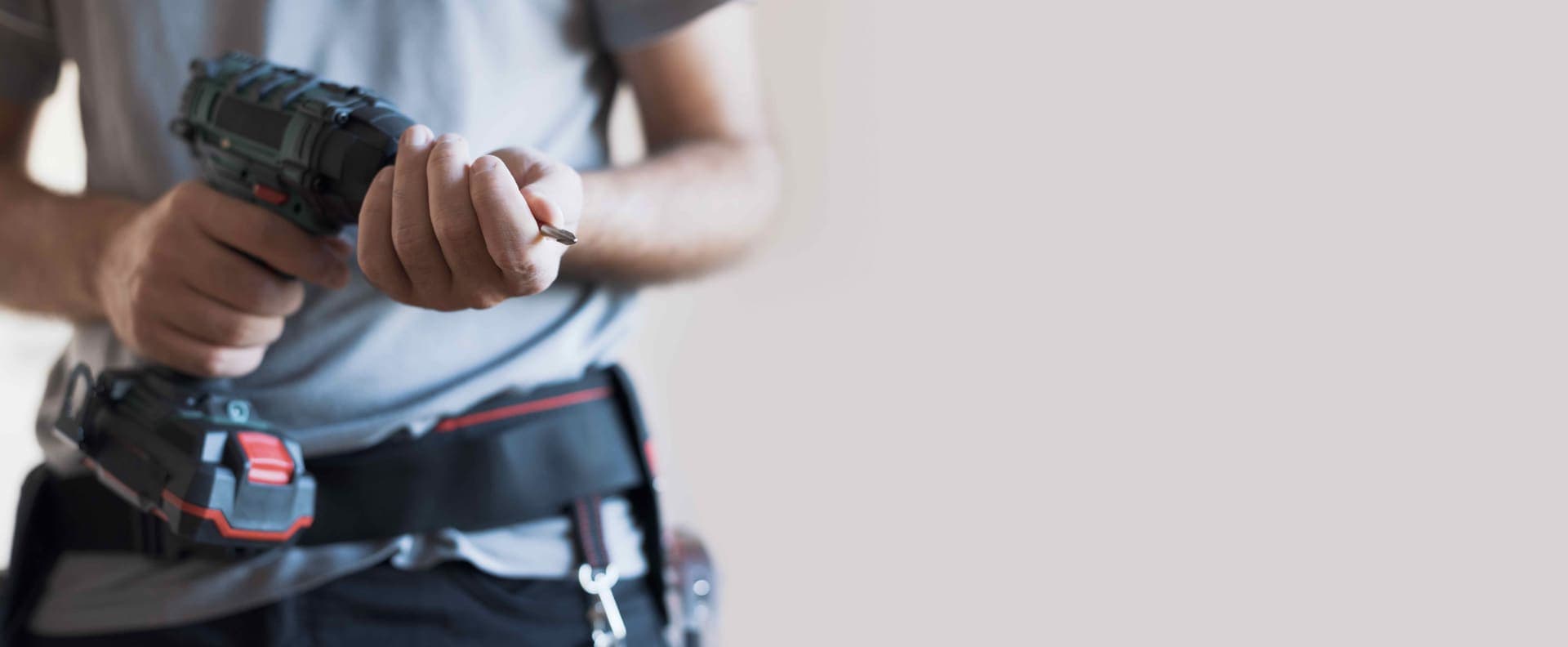 Close-up of a person wearing a tool belt fitting a bit into a cordless drill against a light neutral wall