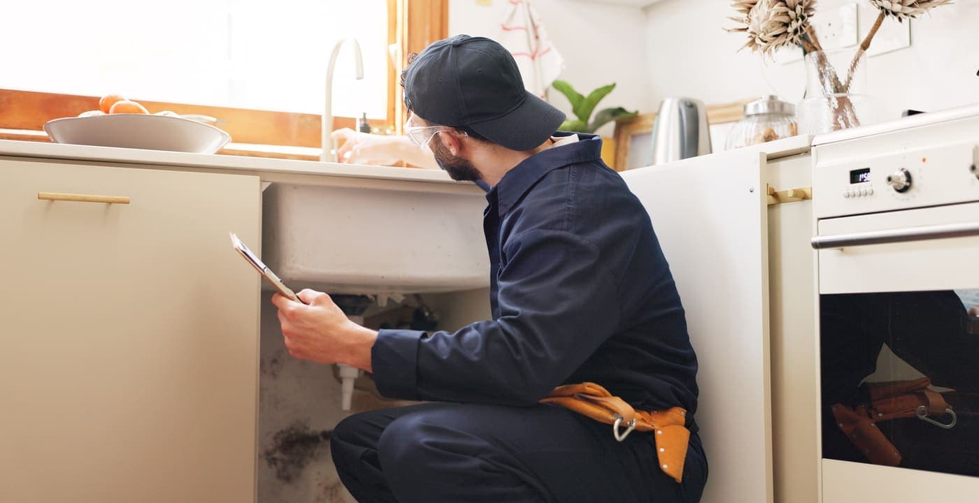 Person in a work cap and safety glasses reviewing a clipboard while checking the plumbing under a kitchen sink