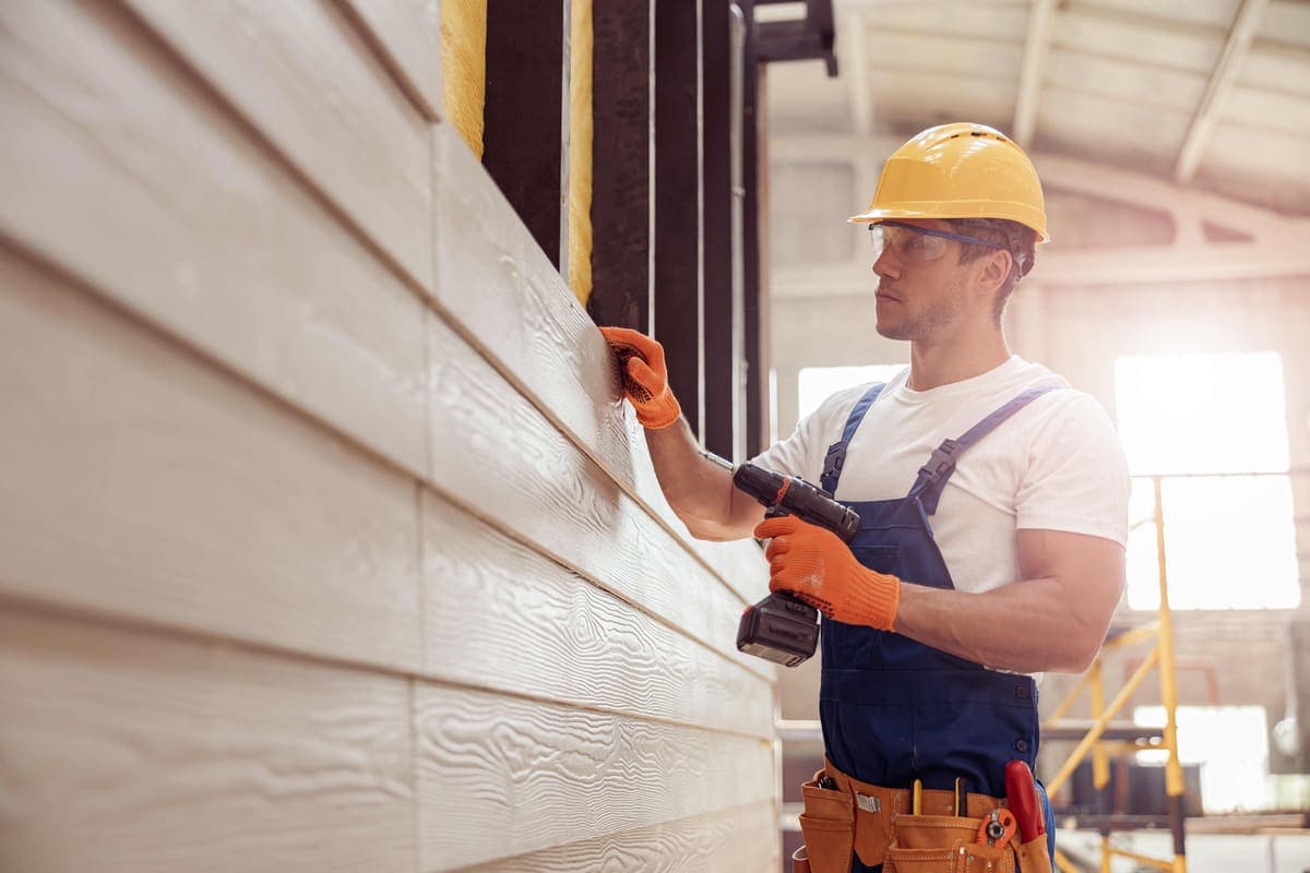 Worker in a hard hat and safety glasses fastening white lap siding panels with a drill