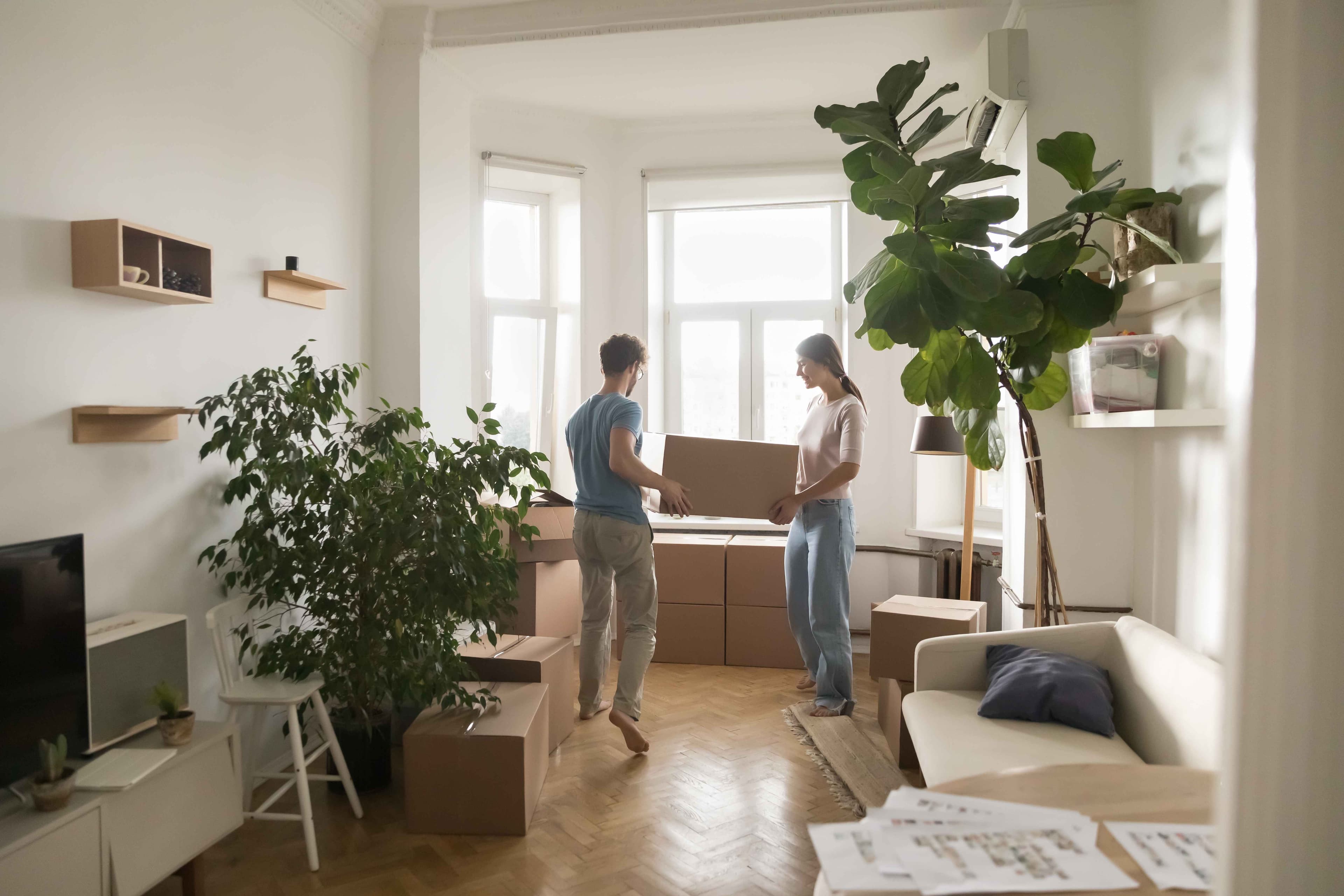 Two people carrying a cardboard box together in an apartment living room surrounded by moving boxes