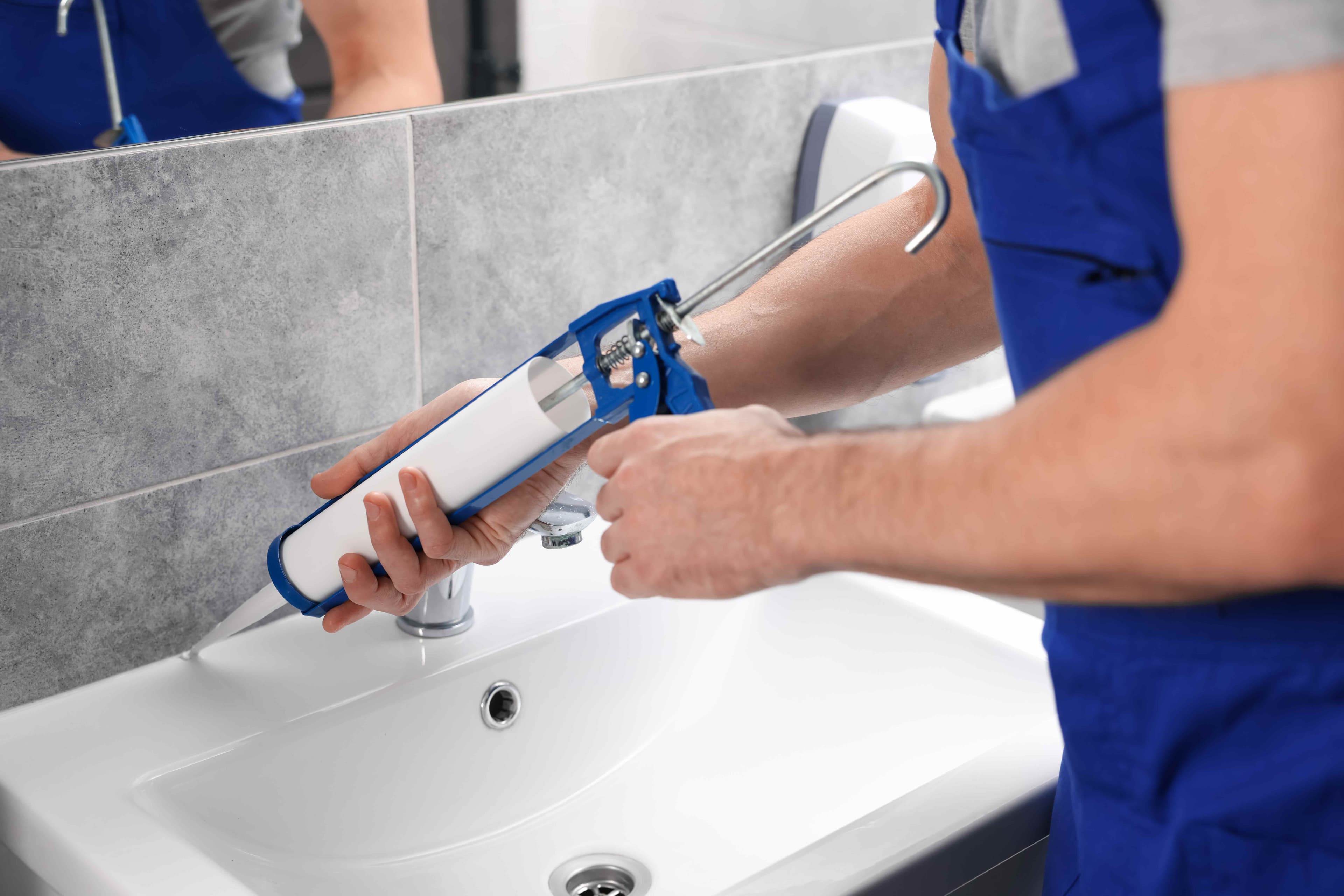 A worker in blue coveralls using a caulk gun to apply sealant around a bathroom sink faucet and tiled wall.
