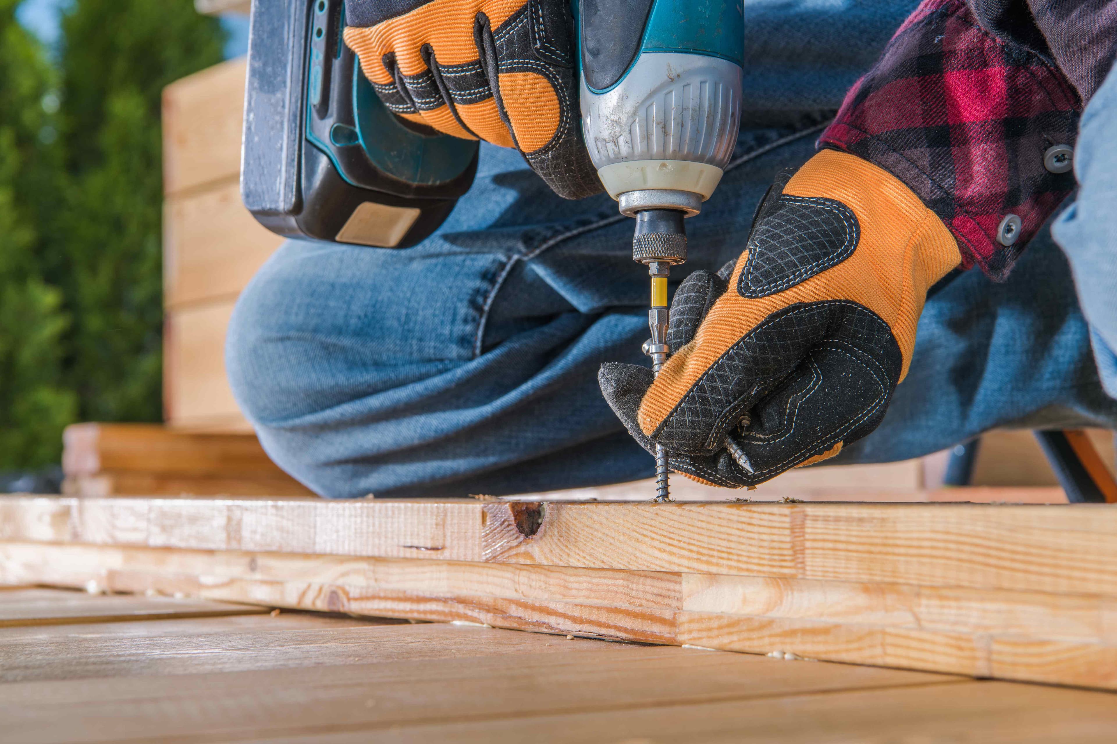 Gloved hands using a cordless drill to drive a screw into wood deck framing near stacked deck boards.