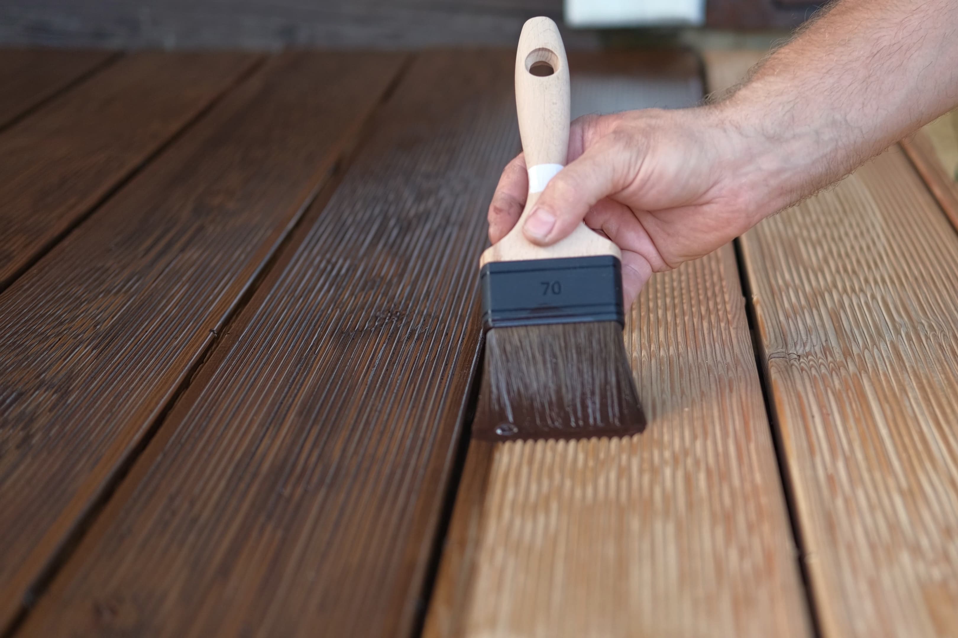 A hand using a paintbrush to apply dark wood stain to deck boards, showing the contrast between stained and unstained wood.