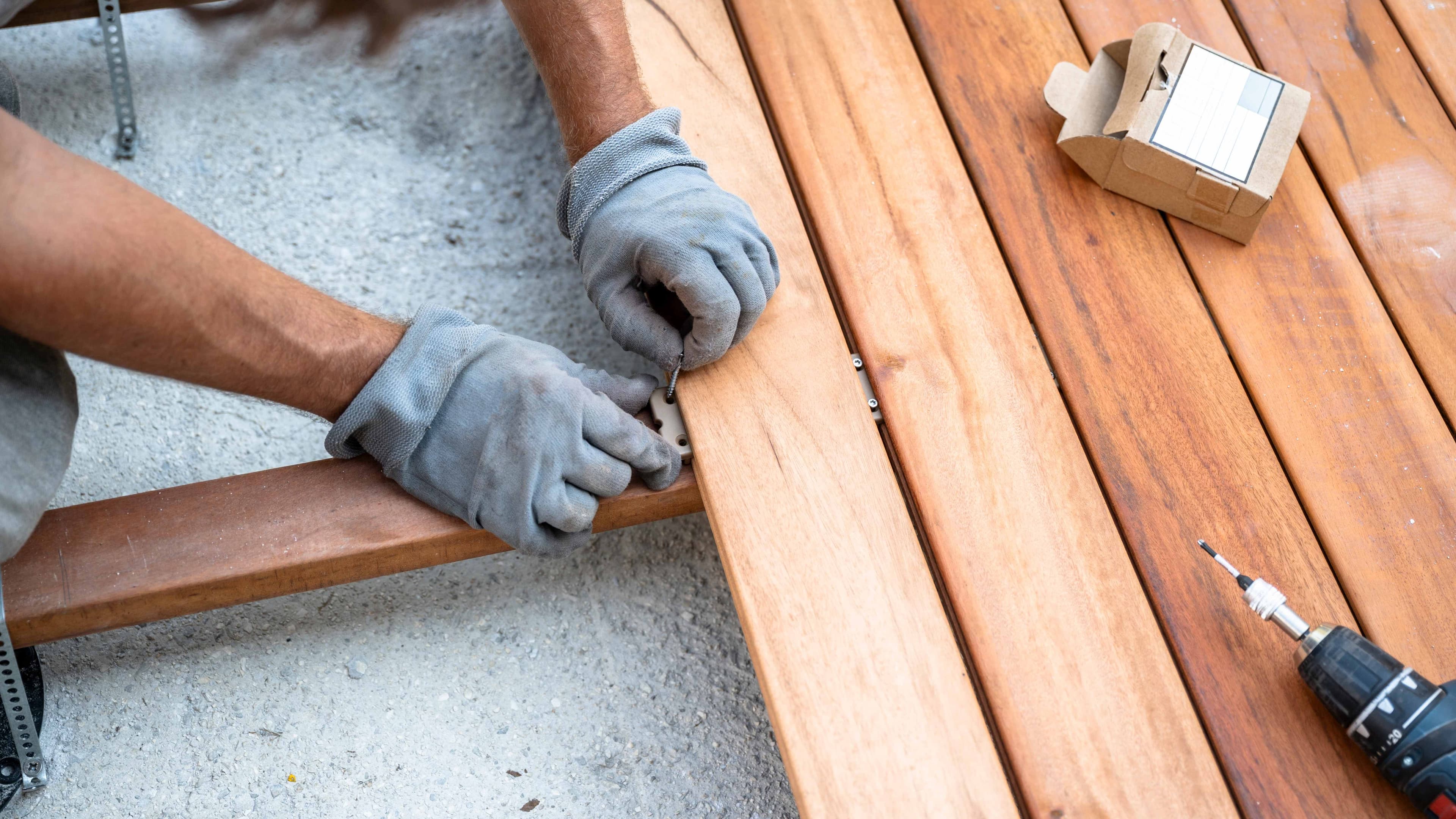 Overhead view of hands installing wood deck boards using hidden fastener clips, with a drill and screw box on the deck surface.