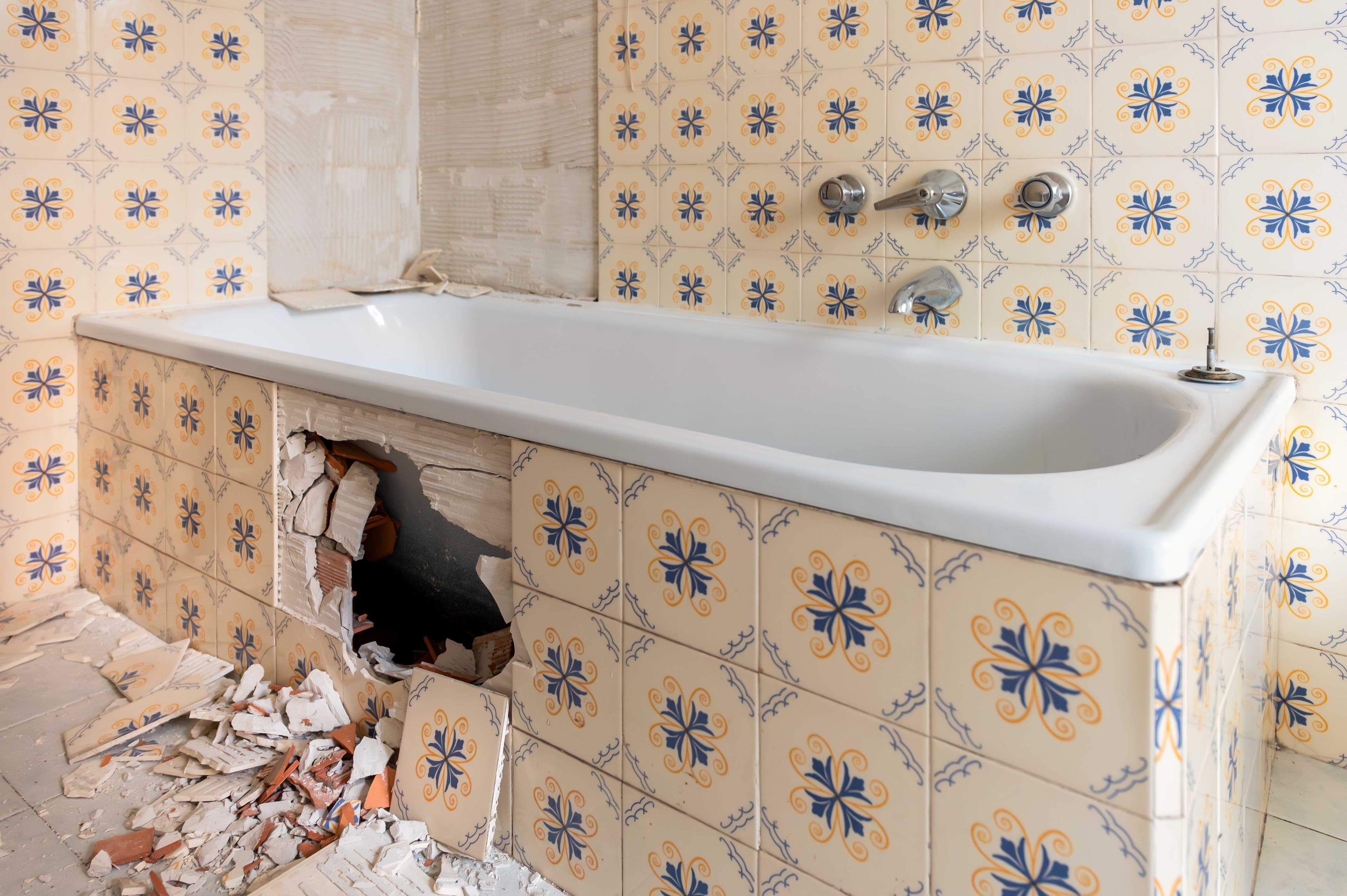 Tiled bathtub surround with a section of tile removed, exposing the wall cavity behind it, with broken tile pieces on the floor