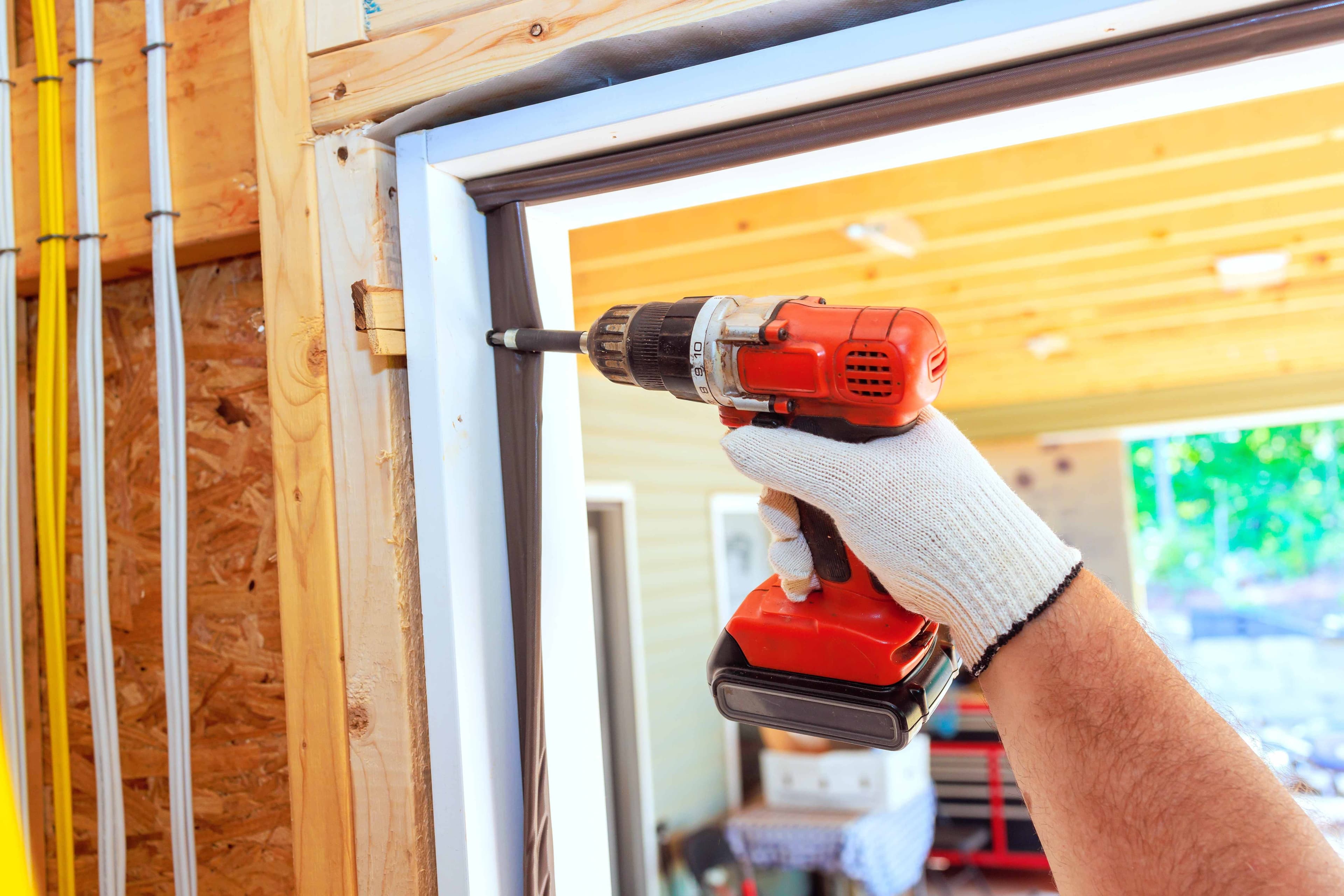 A gloved hand using a cordless drill to fasten a door frame with weatherstripping visible along the jamb.