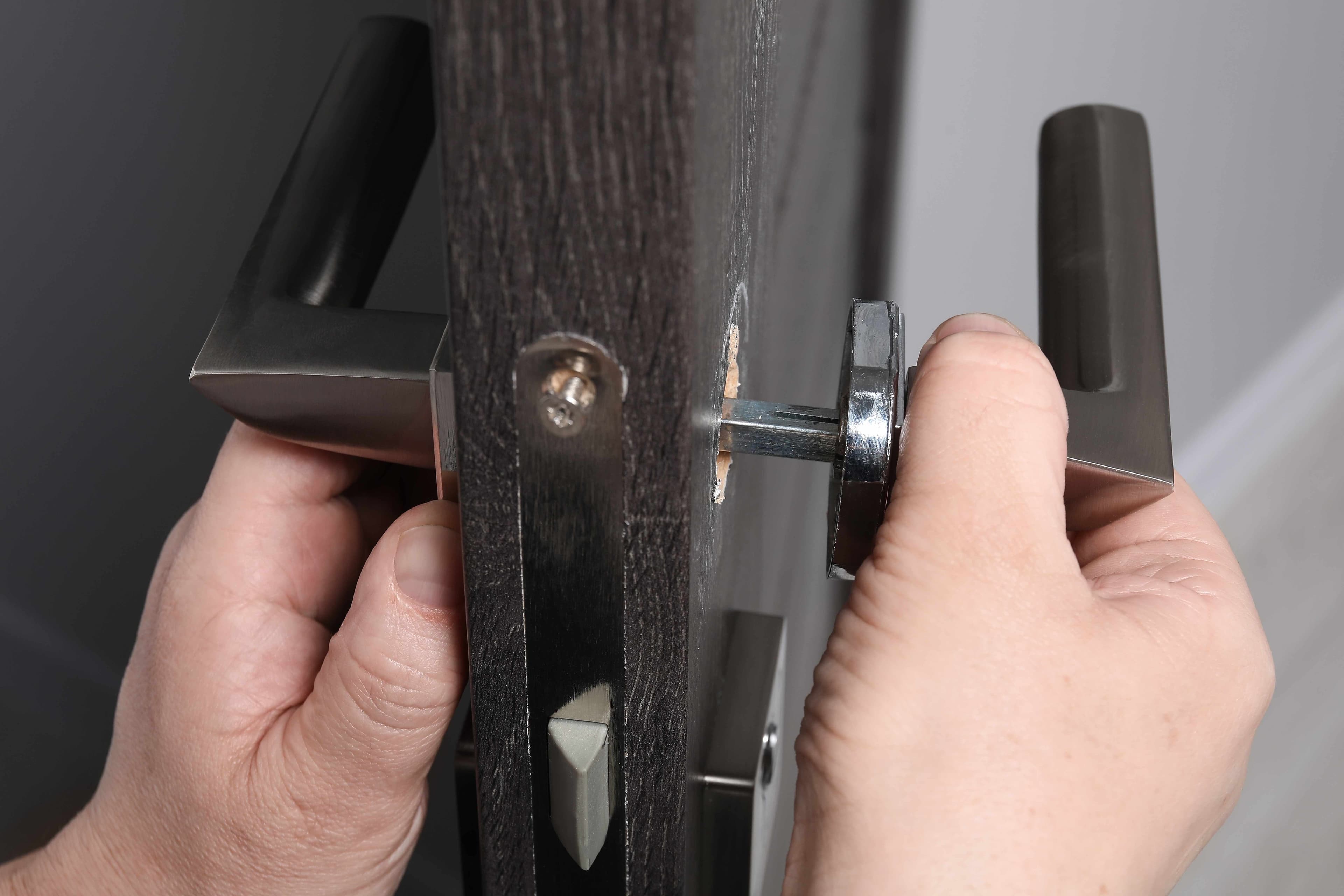 Two hands positioning a lever door handle and hardware on opposite sides of a door edge during installation.