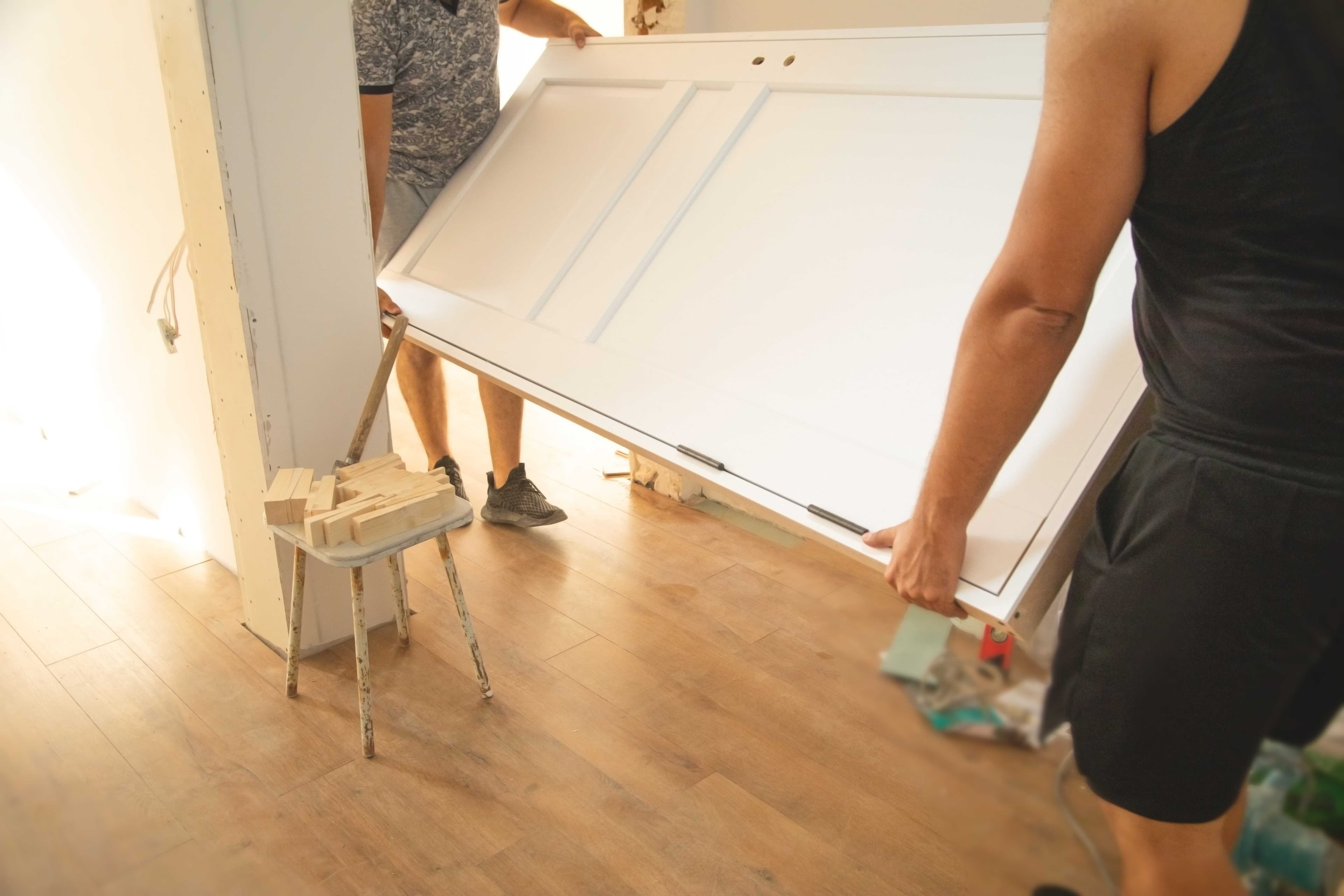 Two people carrying and positioning a white paneled interior door into a framed doorway during installation.