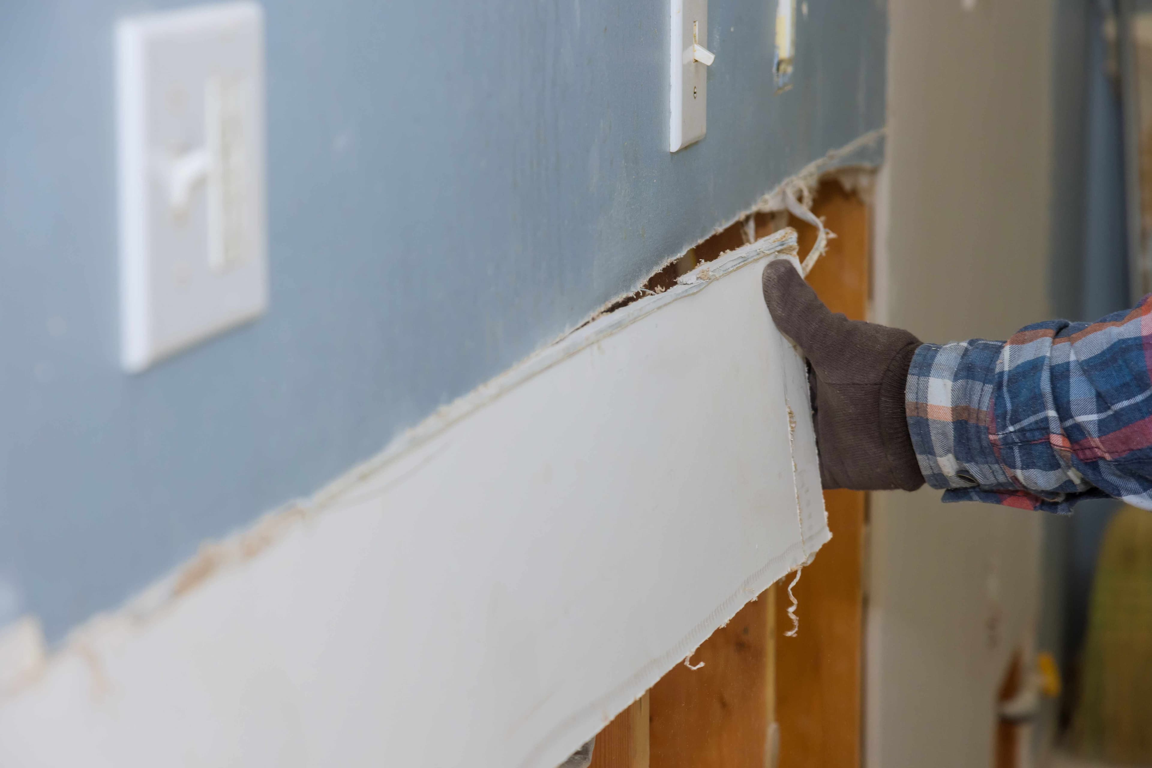 A gloved hand removing a damaged section of drywall near a doorway, exposing the wall framing behind it.