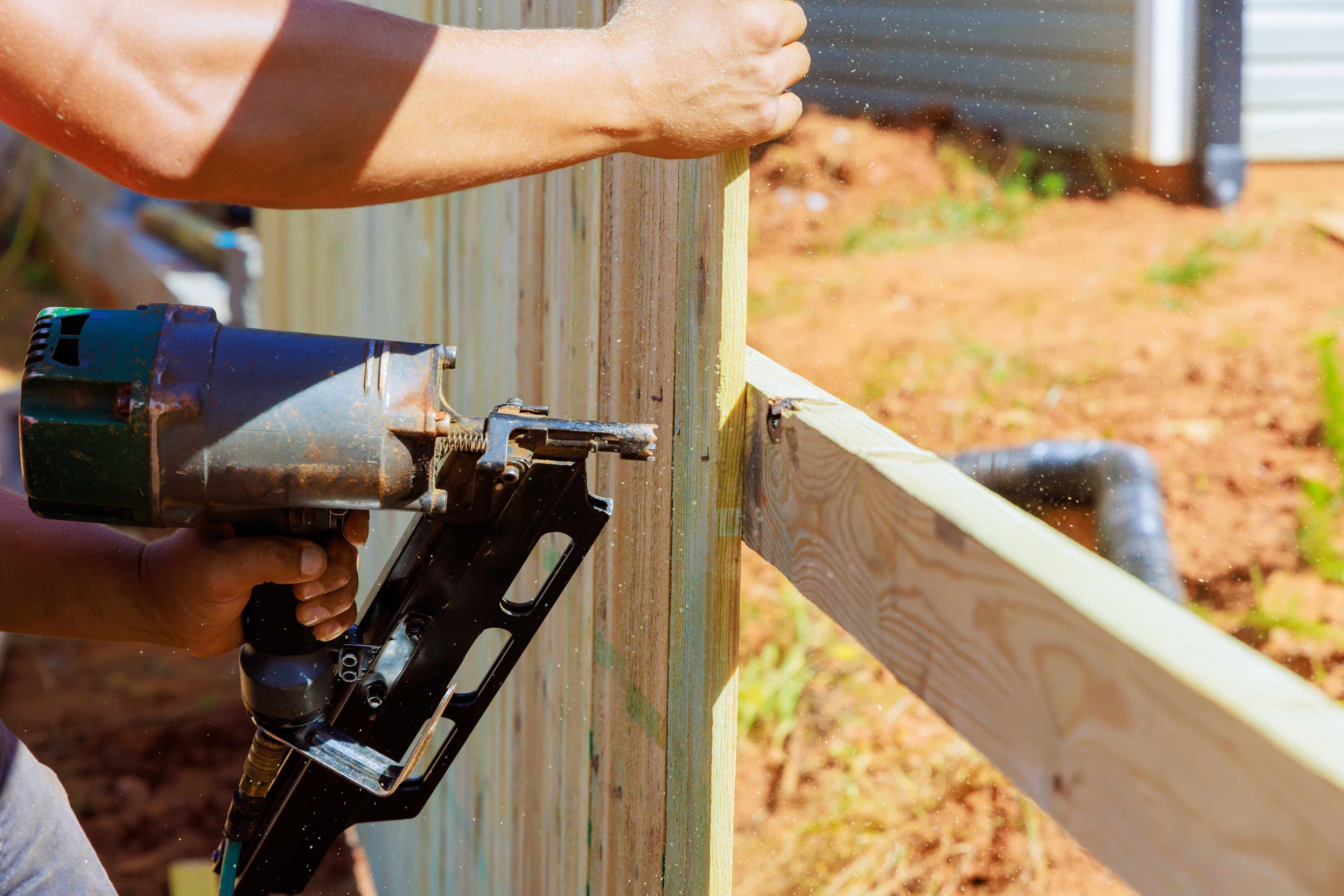 A pneumatic nail gun fastening a horizontal rail board to a wood fence post during fence construction.