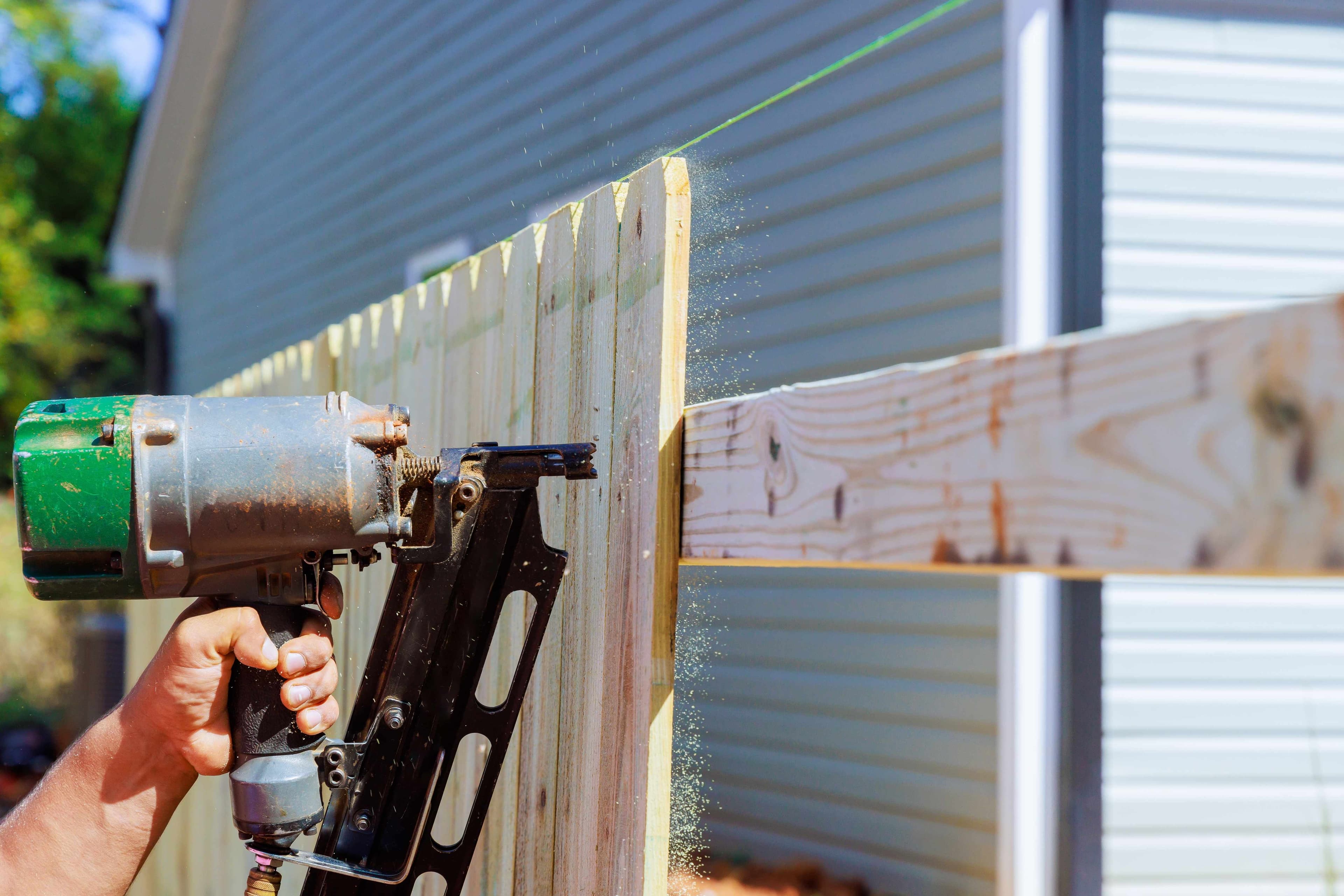 A pneumatic nail gun being used to install wood pickets along a new fence line, with sawdust visible in the air.