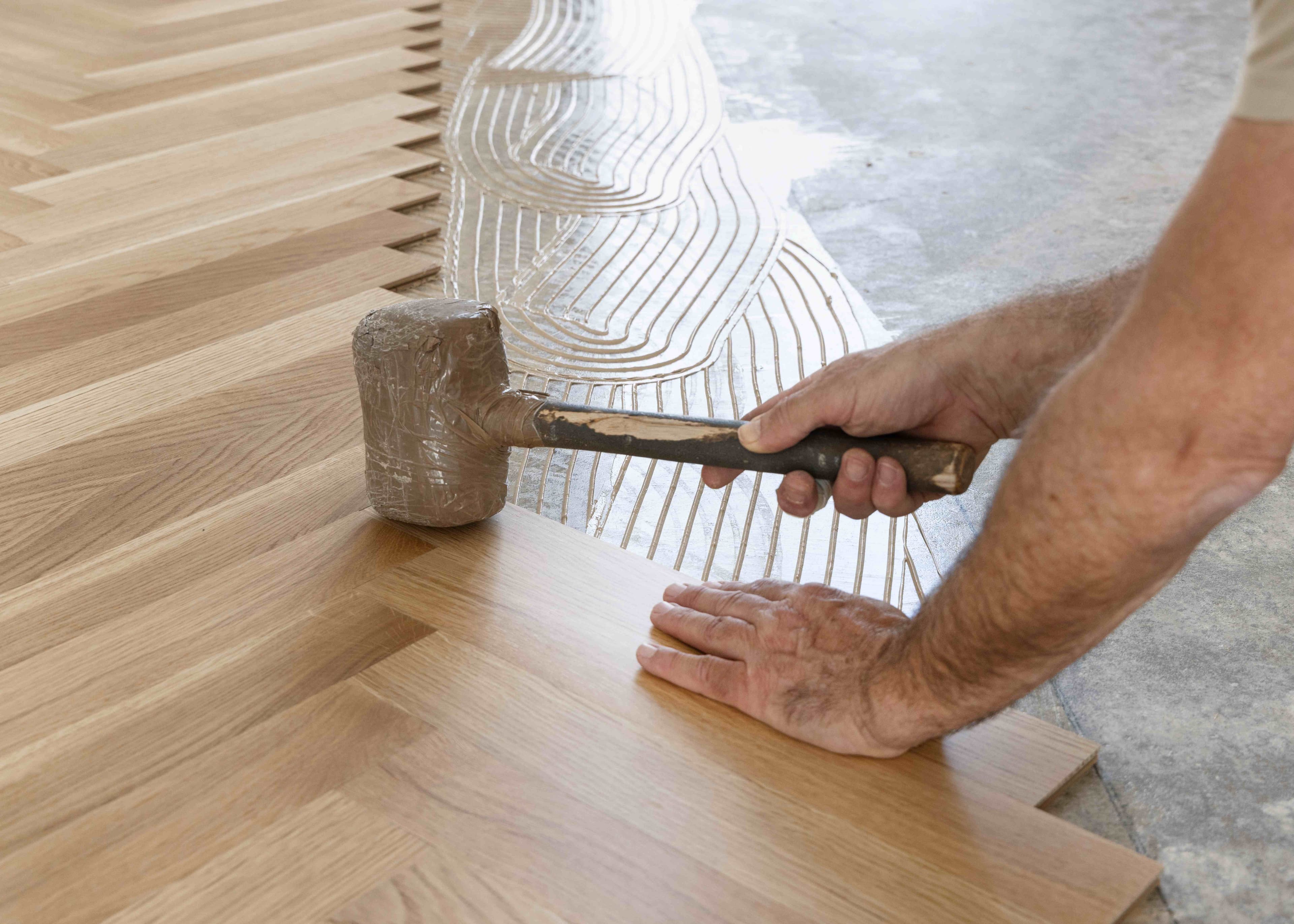 A worker setting a wood plank into adhesive with a rubber mallet during a herringbone-pattern flooring installation.