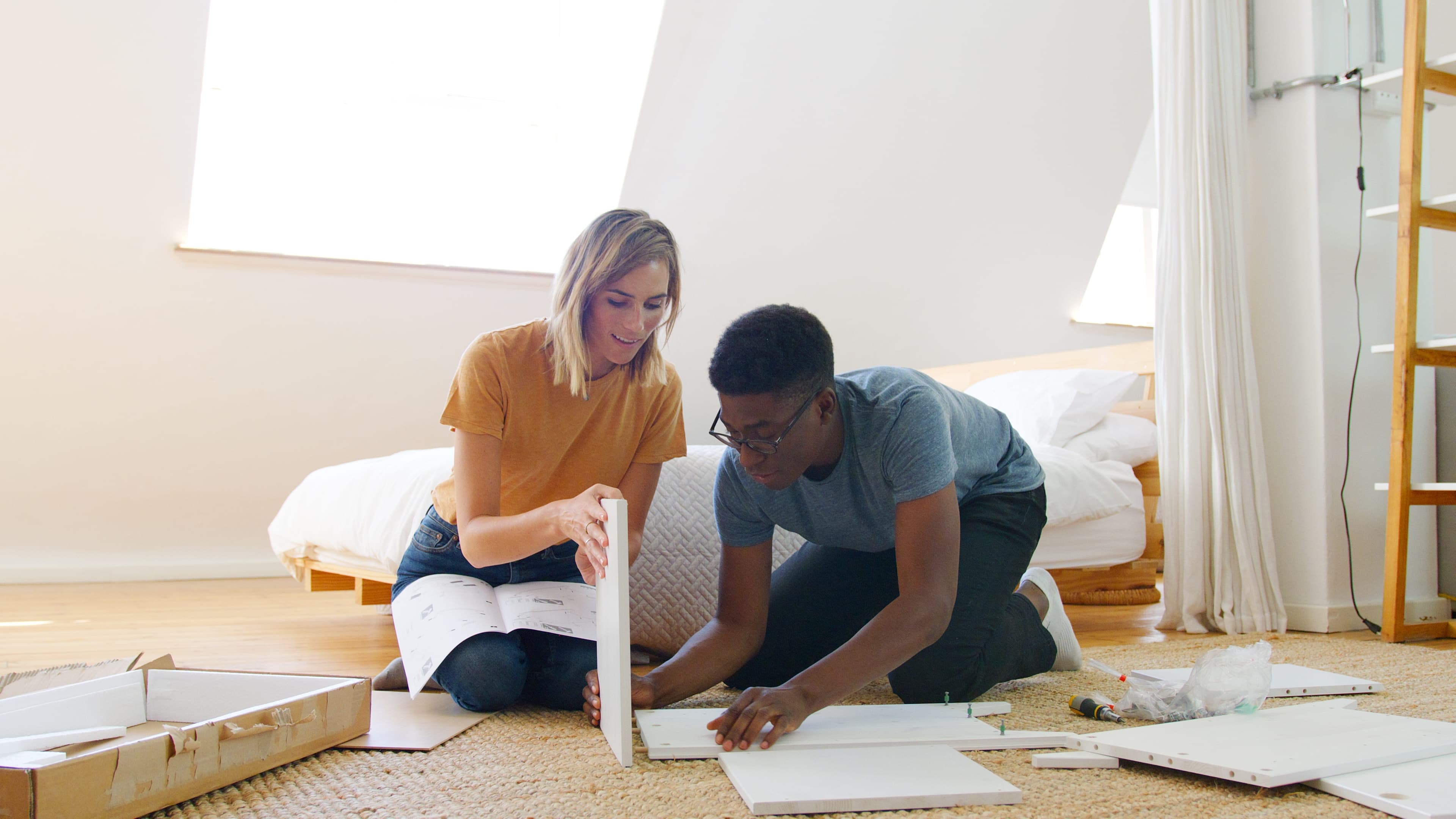 Man and woman assembling white furniture in a bright bedroom, woman holding instruction manual while man works on the furniture