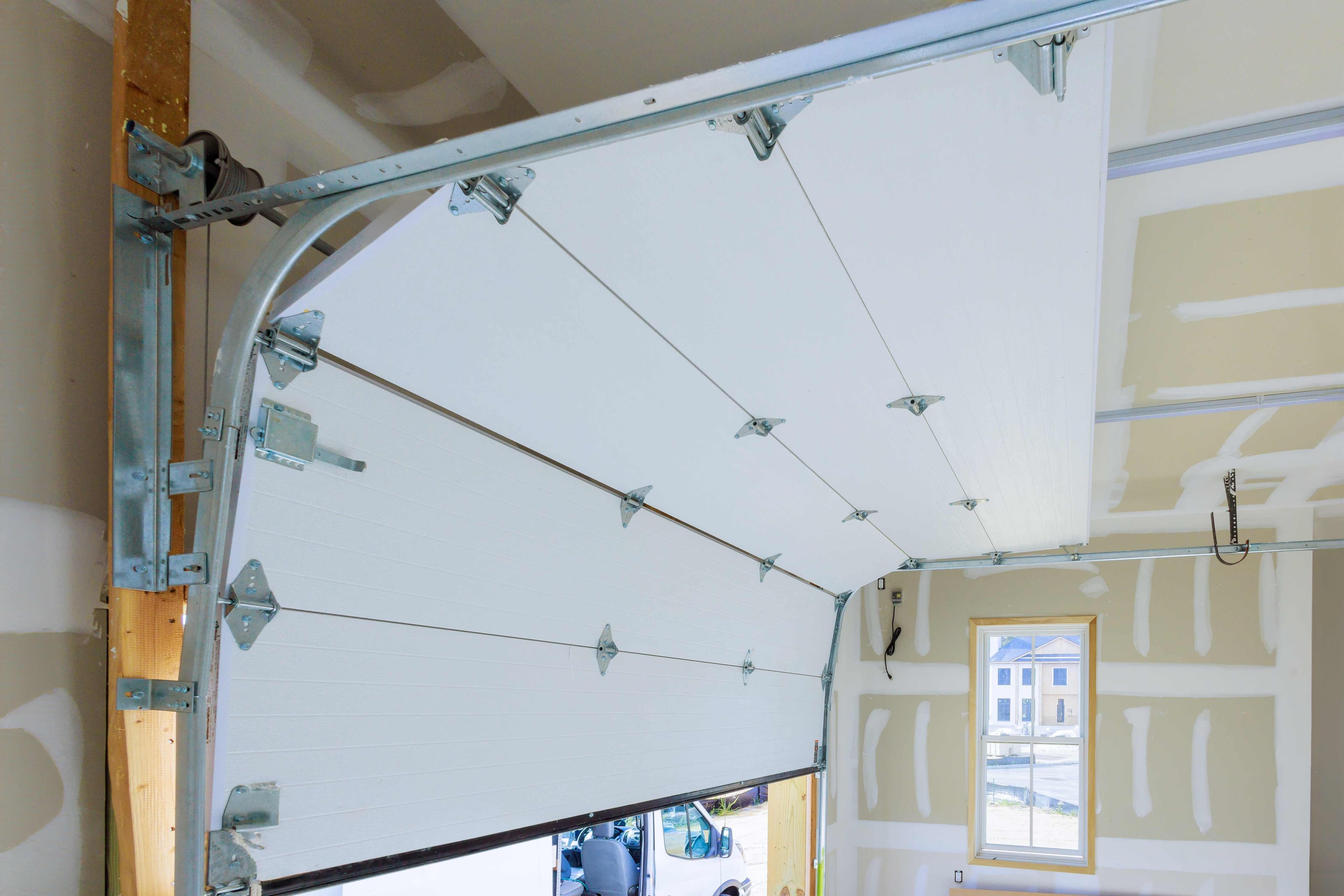 An underside view of a garage door showing door panels, hinges, cables, and the spring assembly along the track.