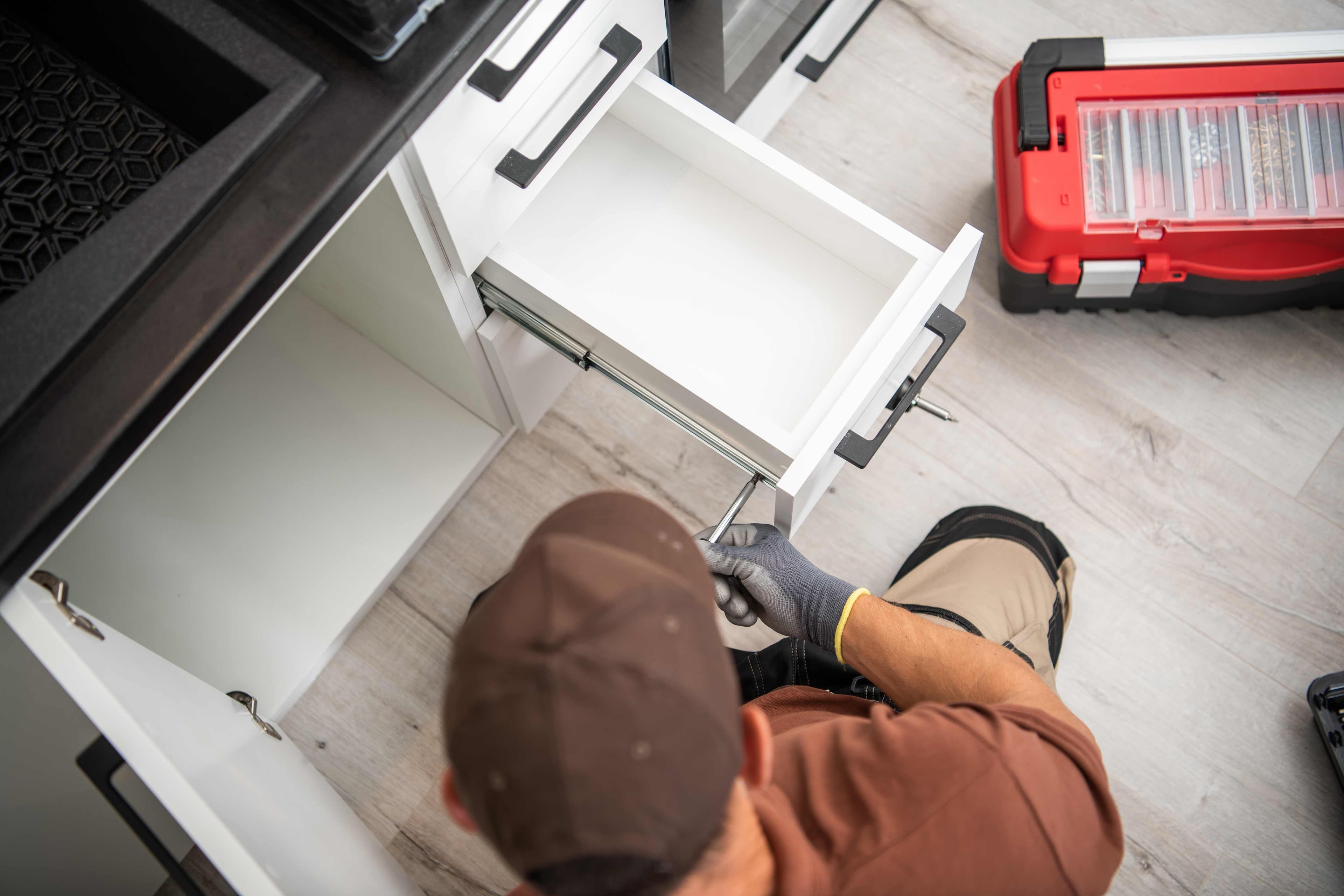 A technician kneeling to adjust a cabinet drawer slide with a screwdriver inside an open kitchen cabinet.