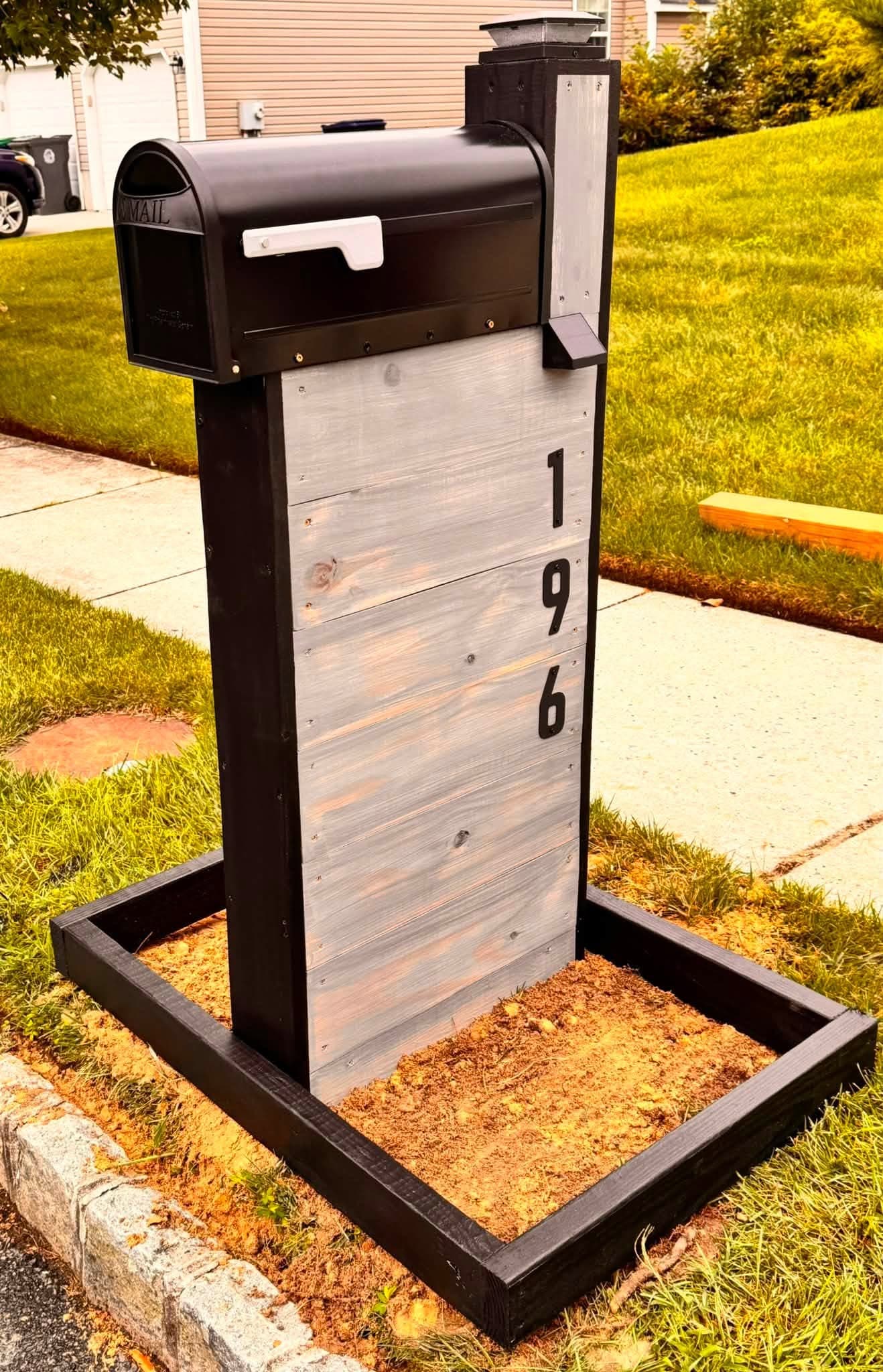 Black residential mailbox mounted on a tall black-and-gray address post within a framed landscape bed beside a sidewalk