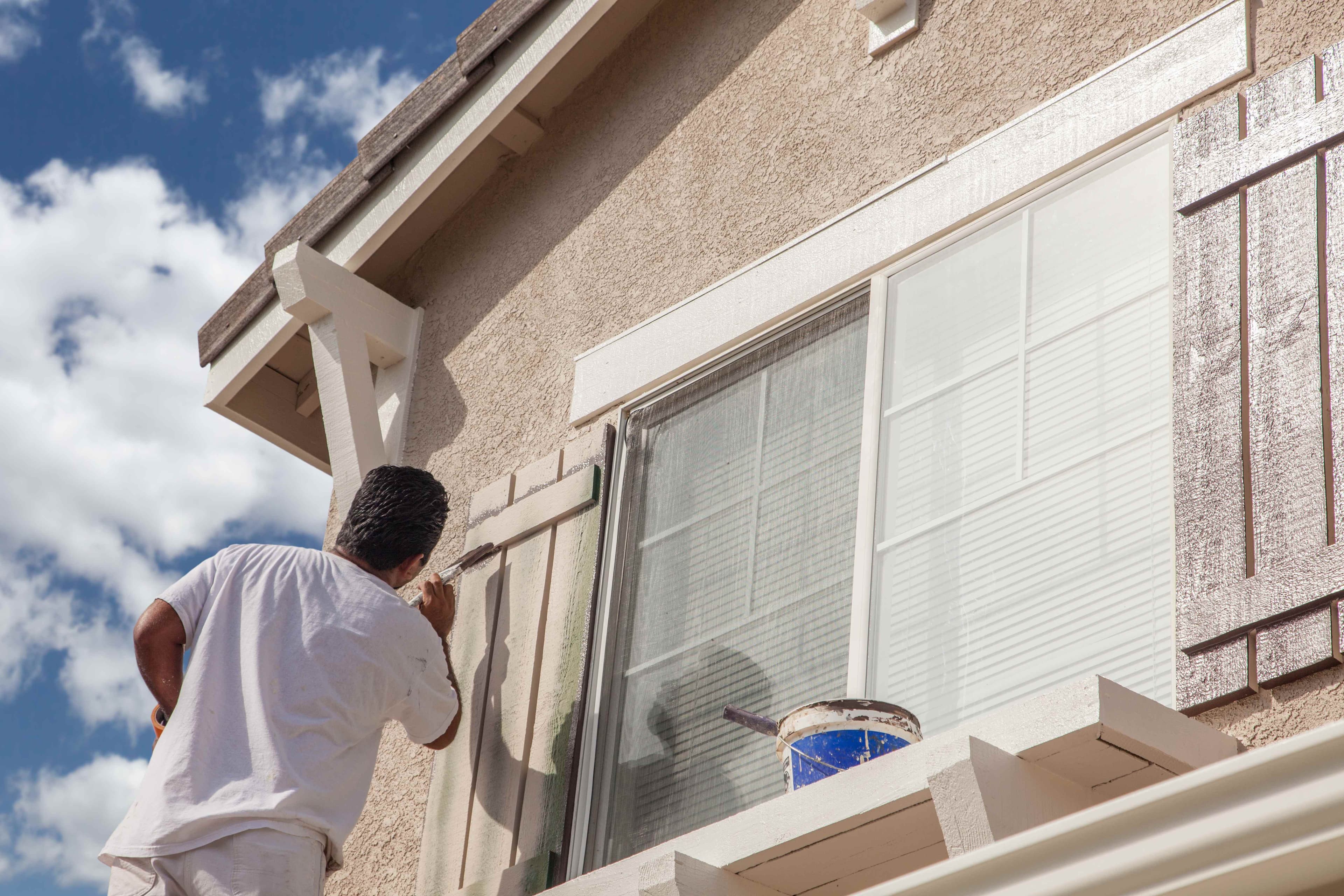 A painter applying paint to exterior window trim and shutters on a stucco home exterior.