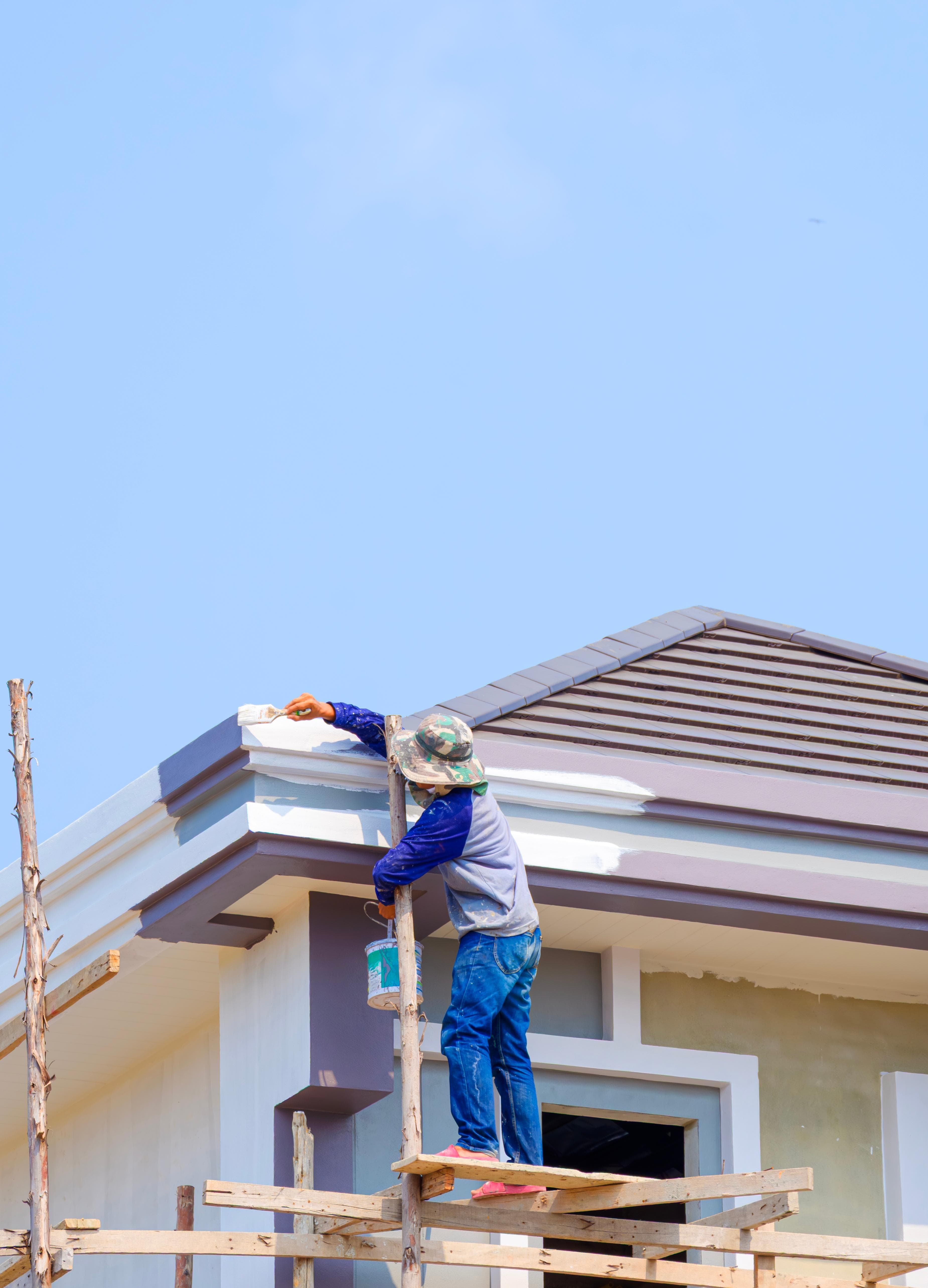 A painter on scaffolding applying paint to the roofline trim of a house exterior with a brush and paint bucket.