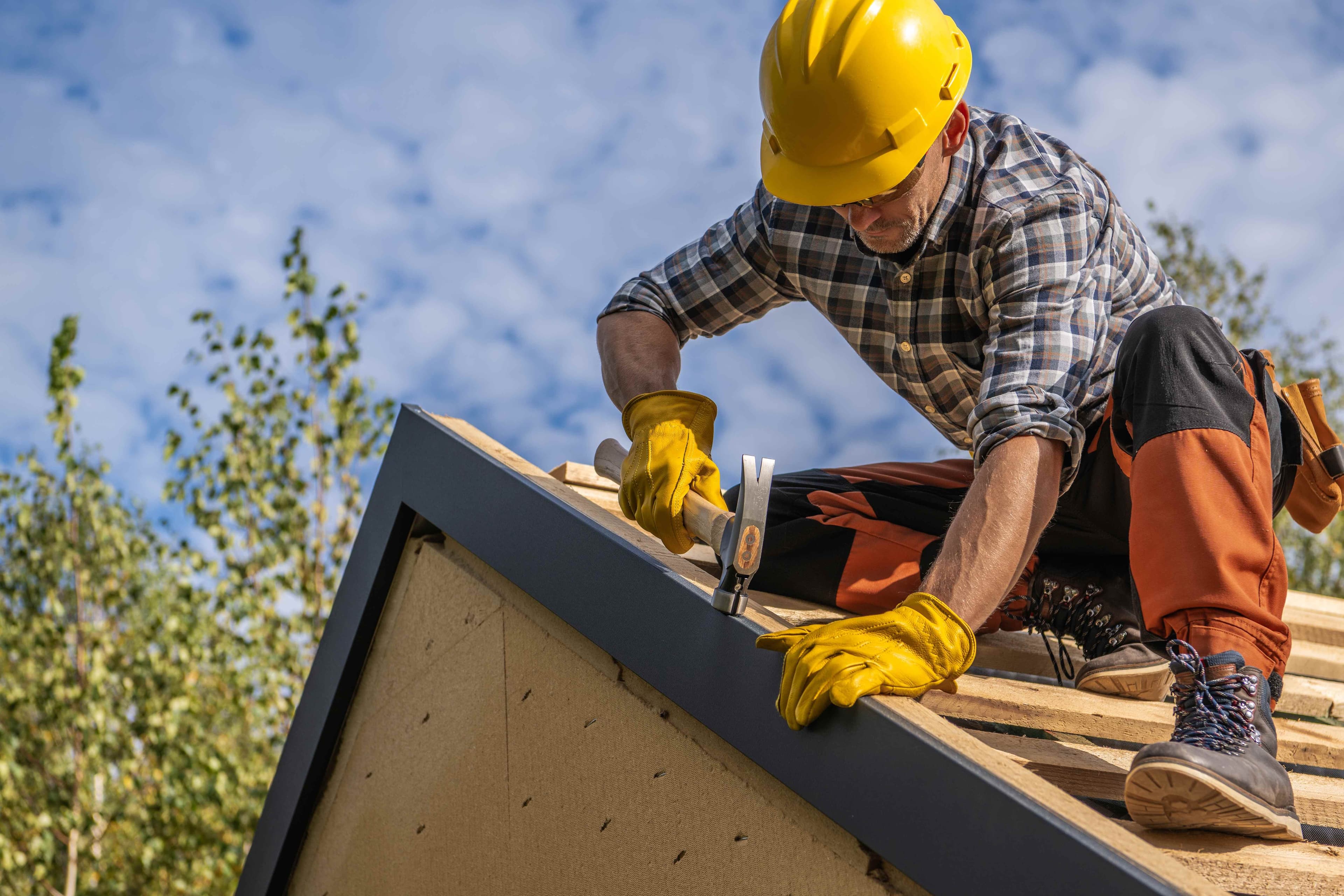 A worker in a hard hat using a hammer to secure metal flashing along a roof edge.