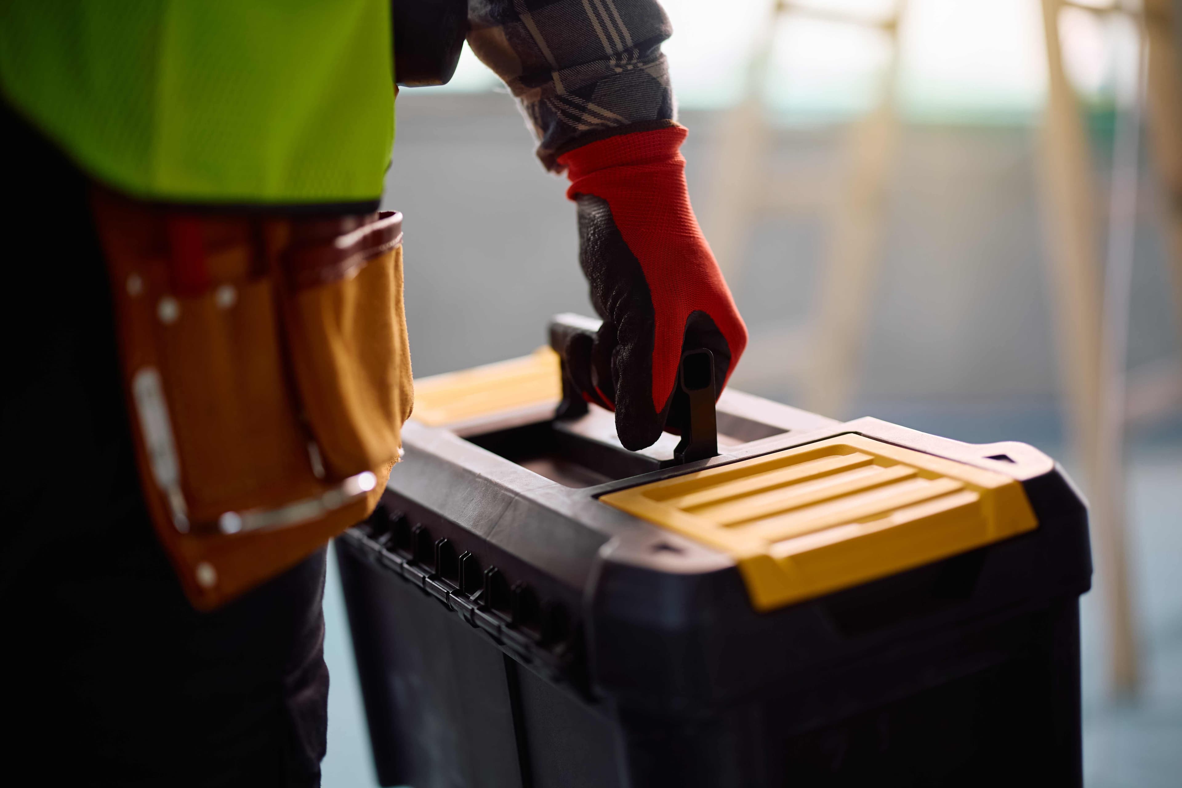 A worker in a hi-vis shirt and tool belt gripping the handle of a black and yellow toolbox on a job site.