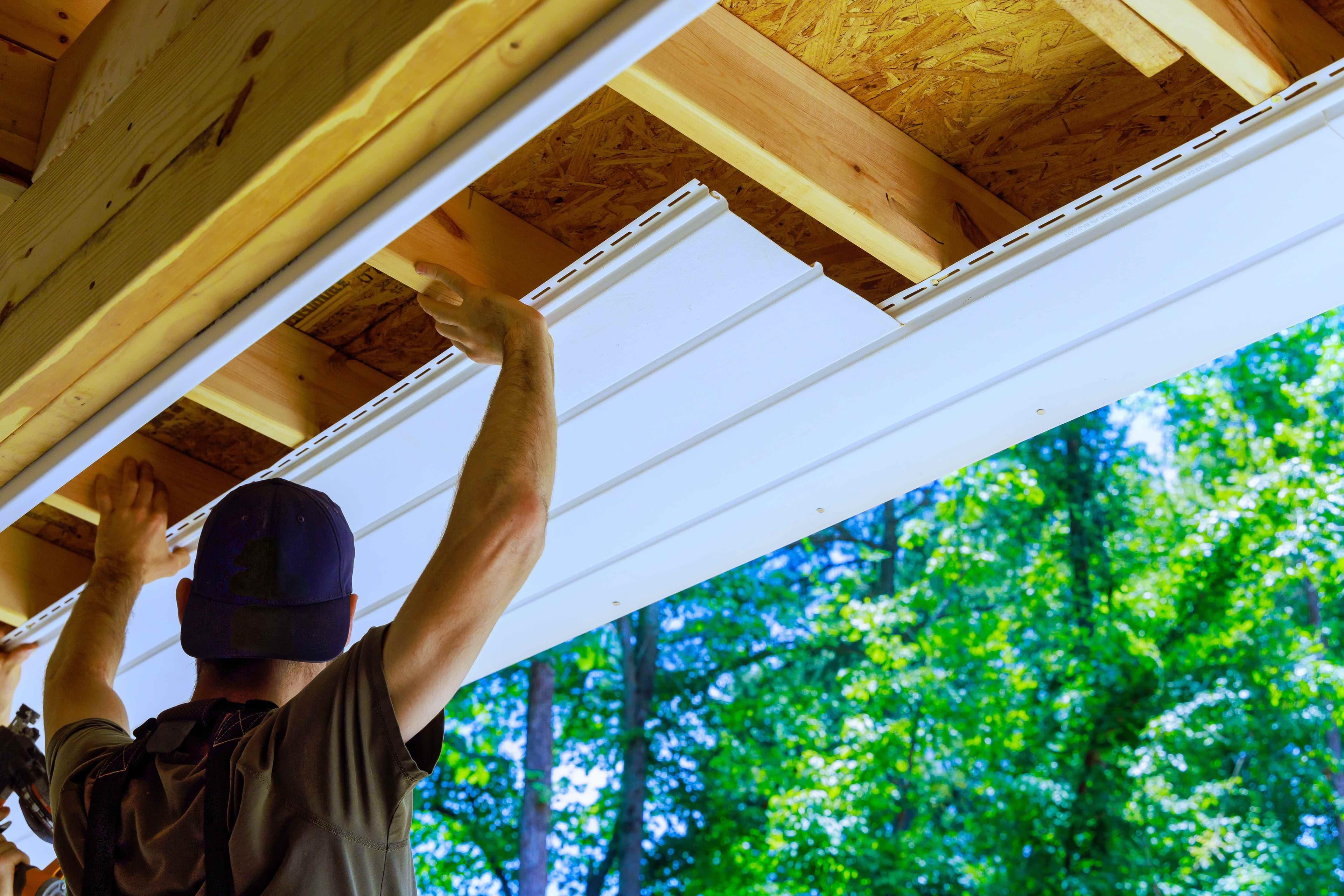 A worker fitting a white vinyl soffit panel into the framing under a roof eave.