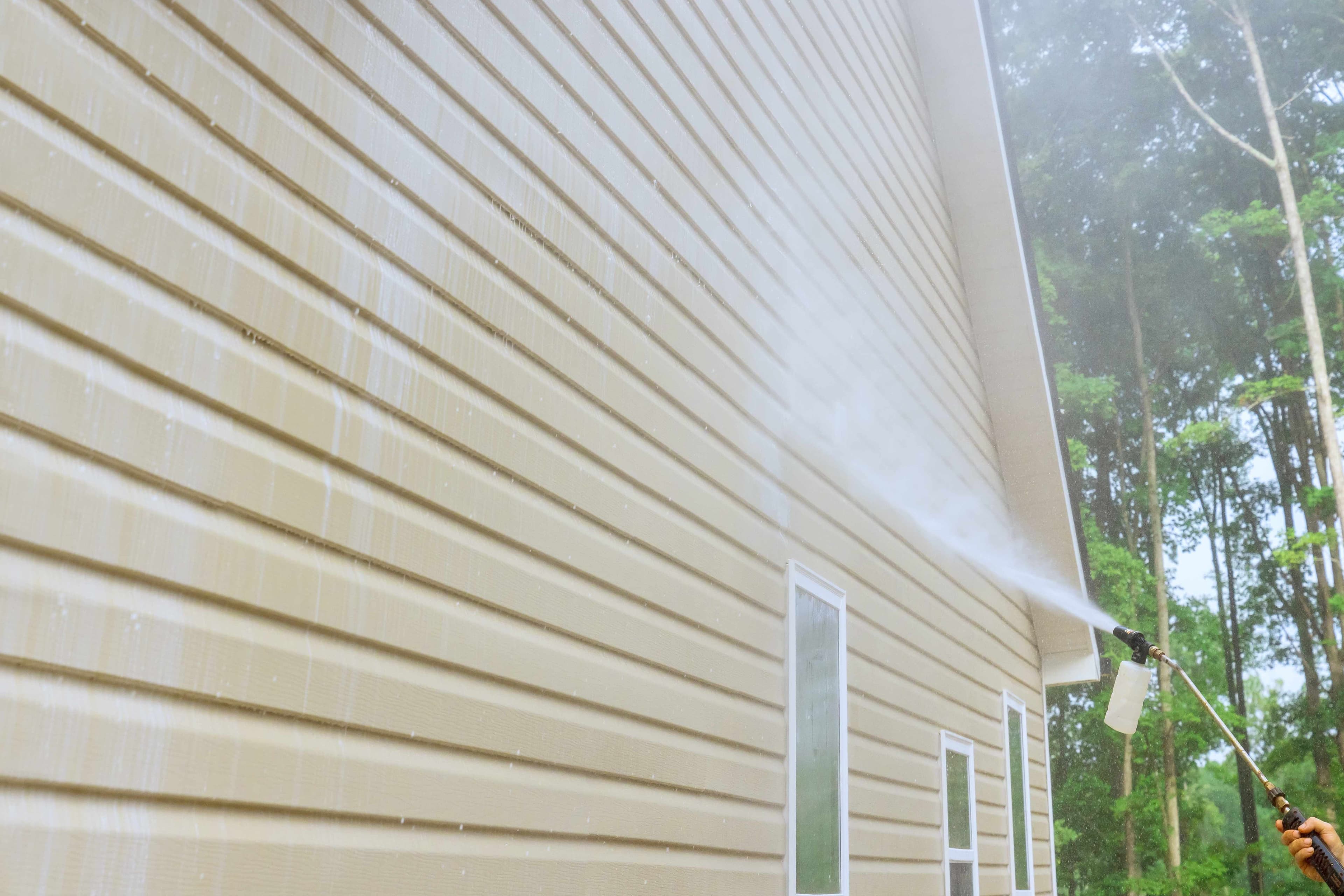 A pressure washer spraying tan vinyl siding on the exterior wall of a house, with window trim visible below the roofline.