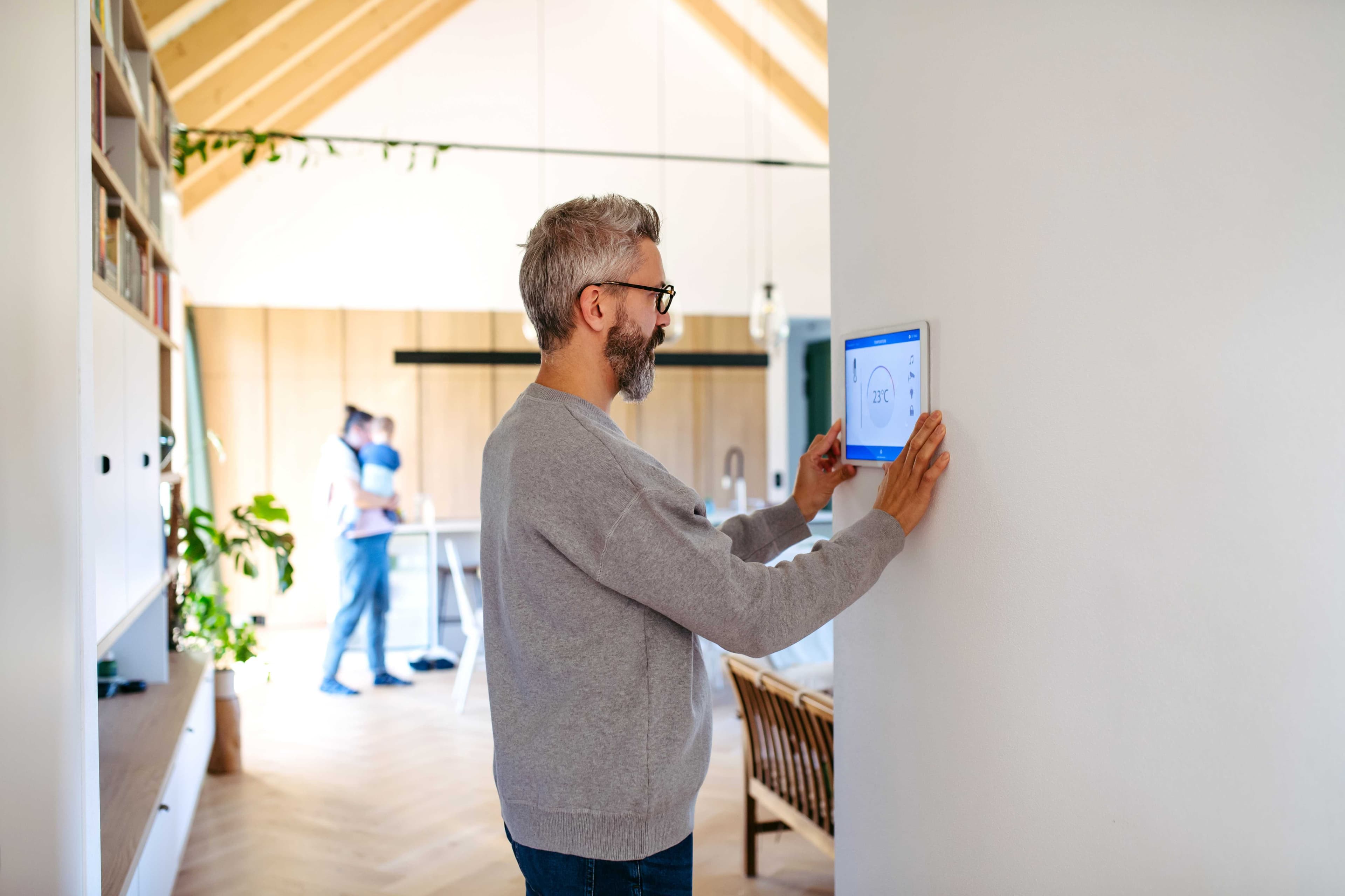 A homeowner adjusting a wall-mounted smart home control panel that displays temperature and device shortcuts.