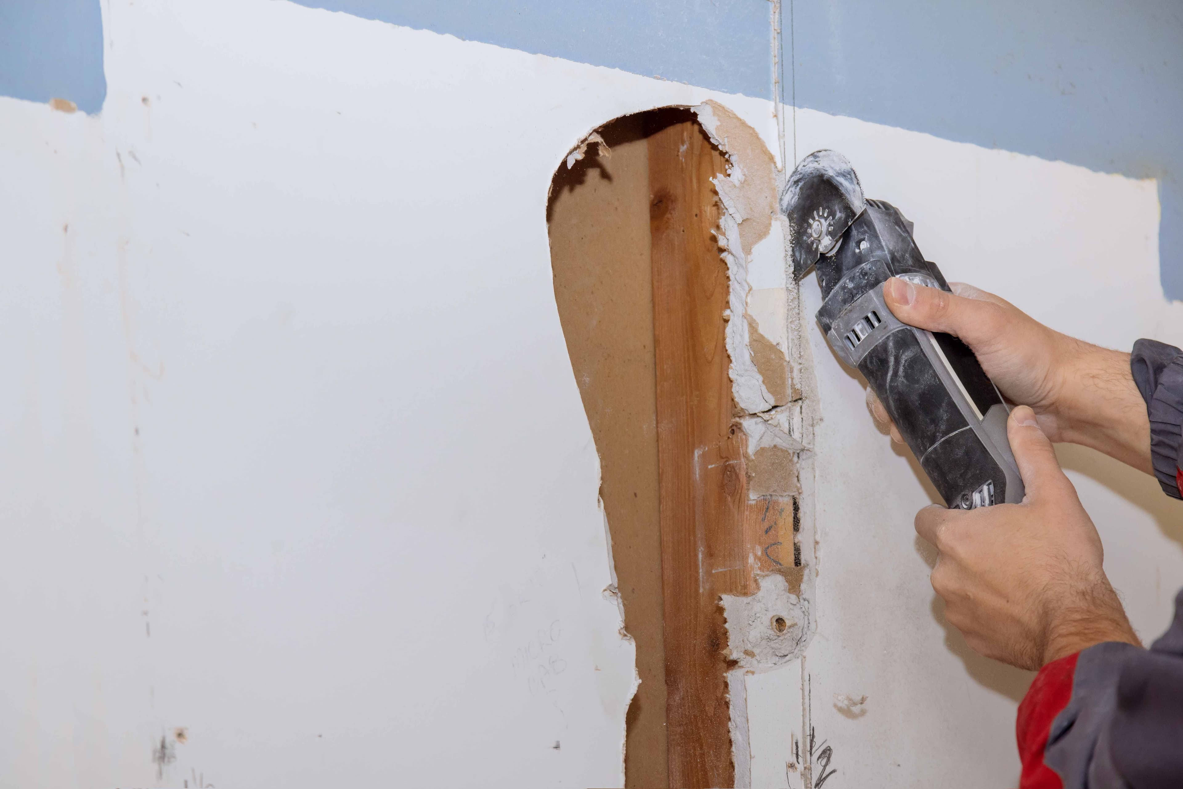 Worker's hands using an oscillating multi-tool to remove and repair water-damaged drywall around wooden studs and wall cavities